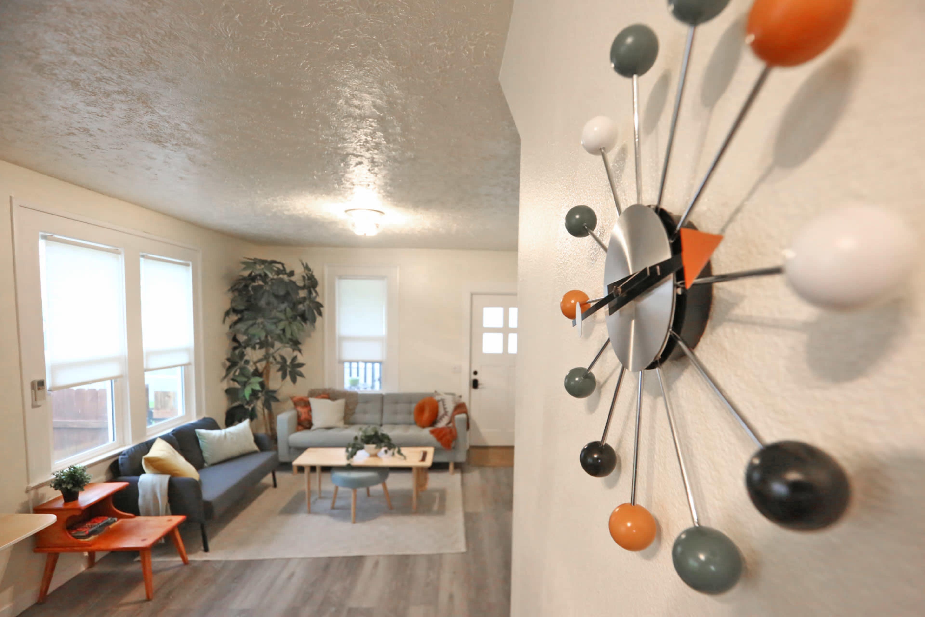 A modern living room features a gray sofa, a coffee table, and a large plant, all seen from an angled perspective that includes a colorful wall clock.