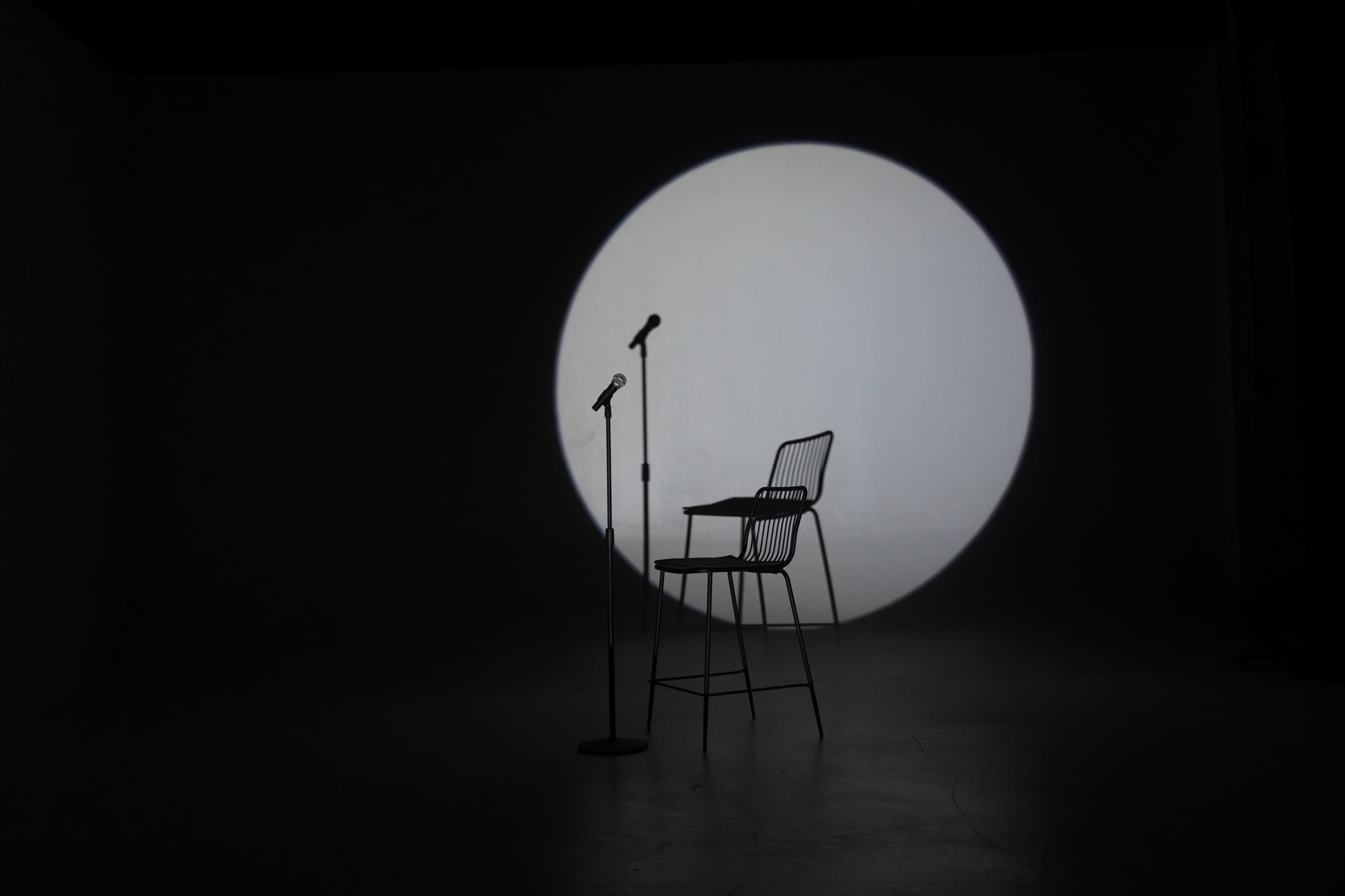 A tall, empty chair is positioned under a spotlight with a microphone beside it, casting a shadow on a plain background.