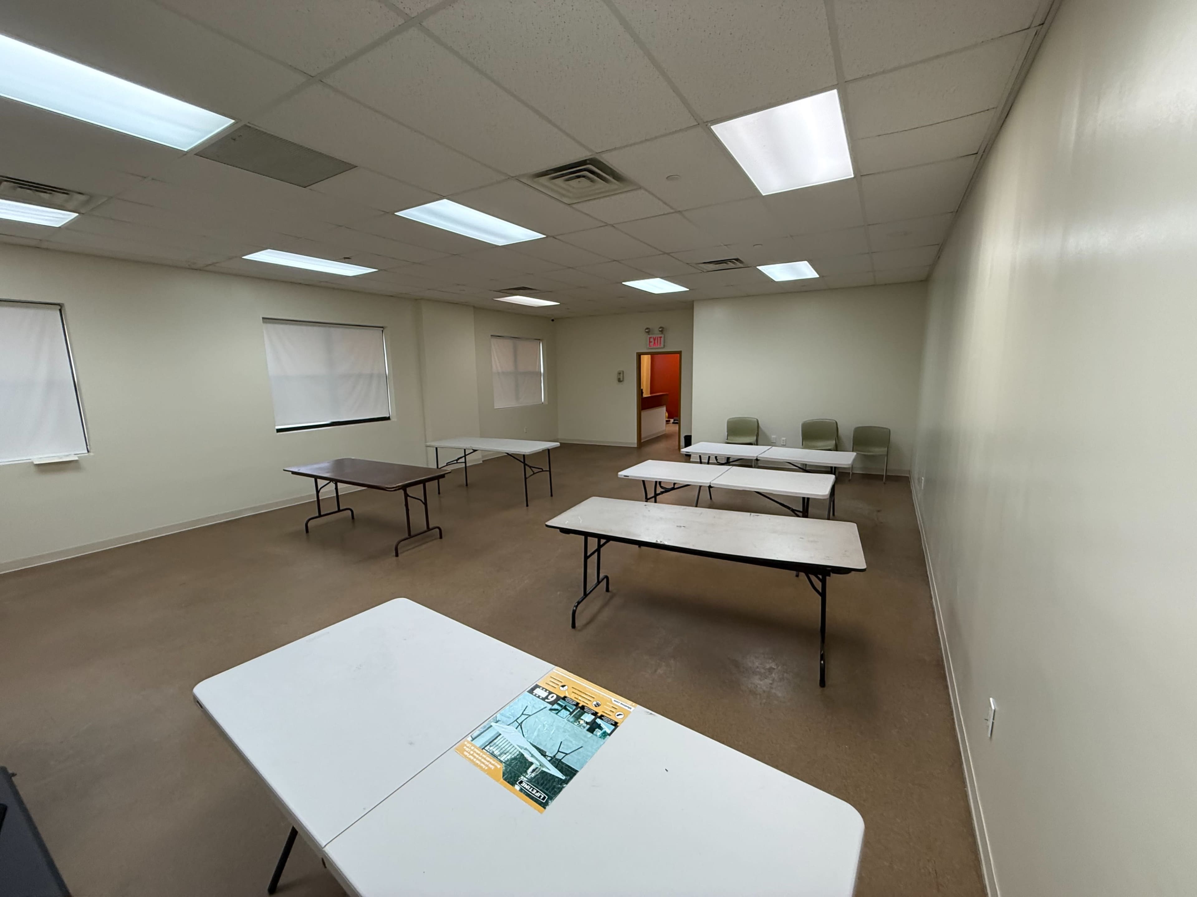 The image shows a spacious room with several tables arranged in a grid pattern and a few chairs against the wall.