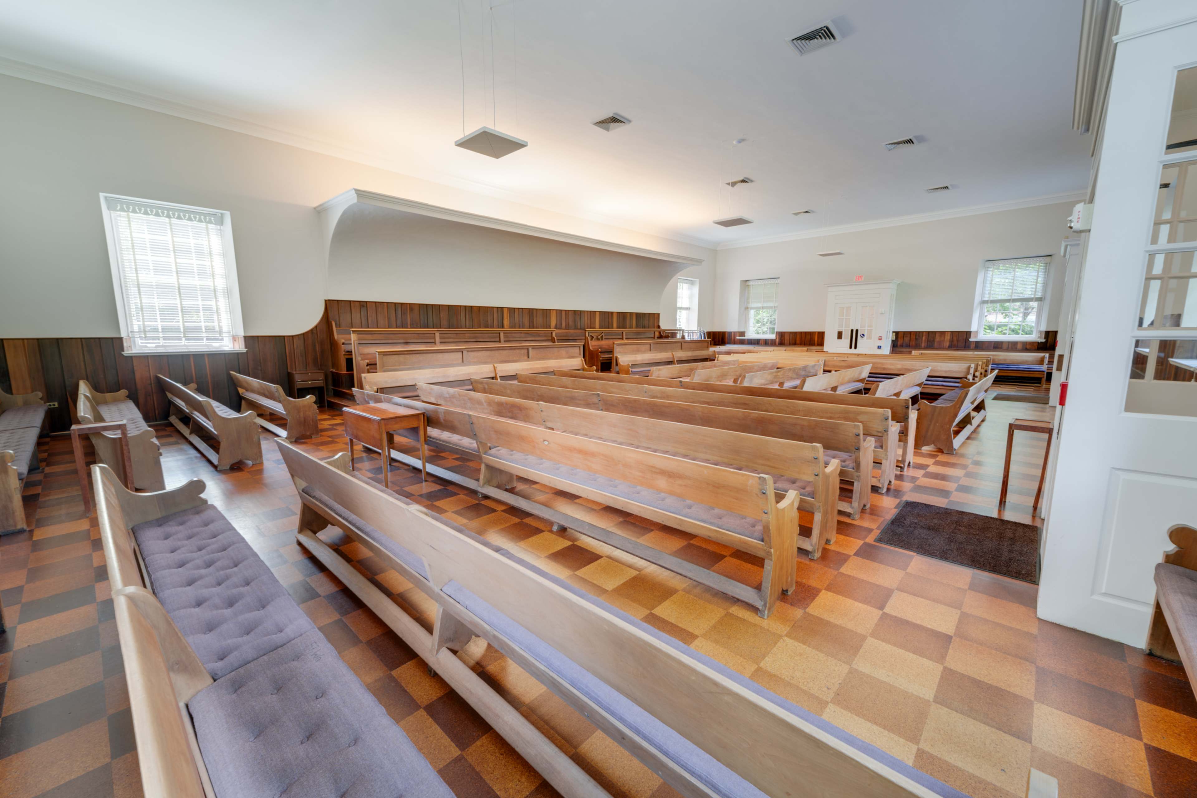 An empty interior of a church with wooden pews arranged in rows and a checkered floor.