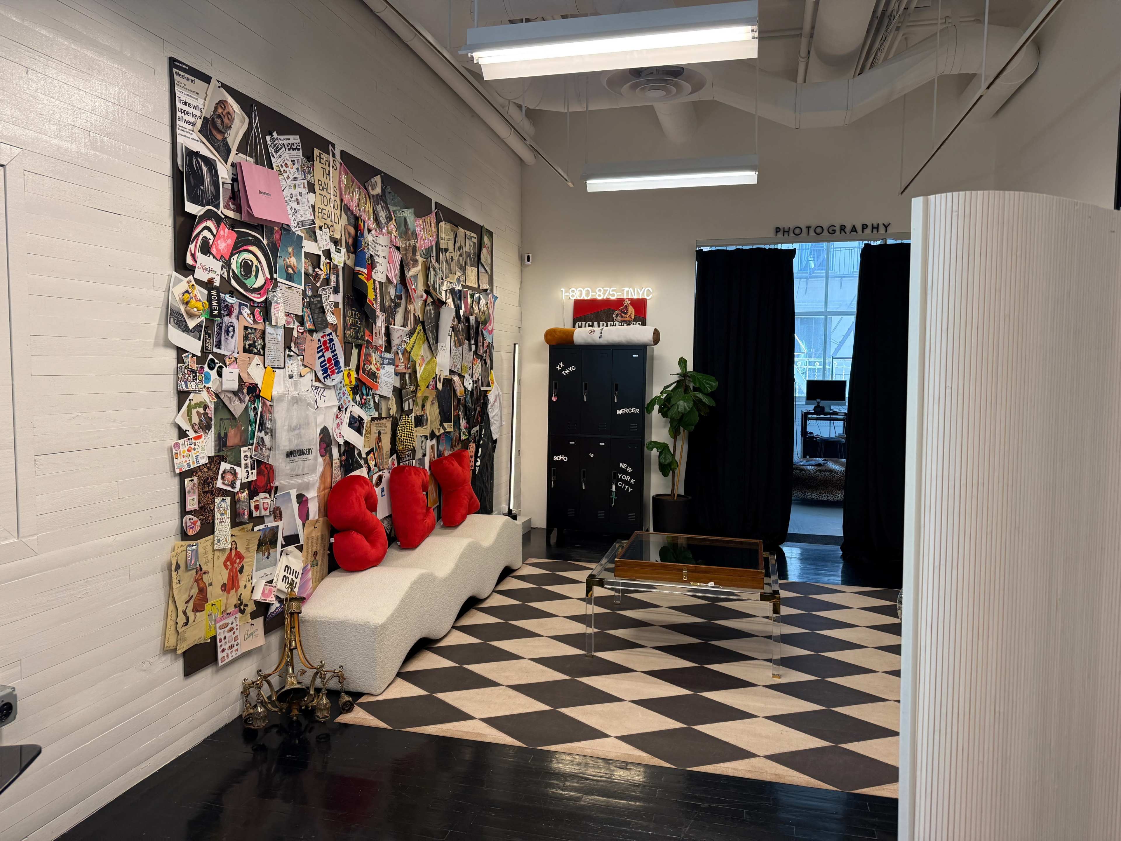 The image shows a room with a black-and-white checkered floor, a white seating area against a wall covered in various papers and images, and dark curtains at the end of the space.