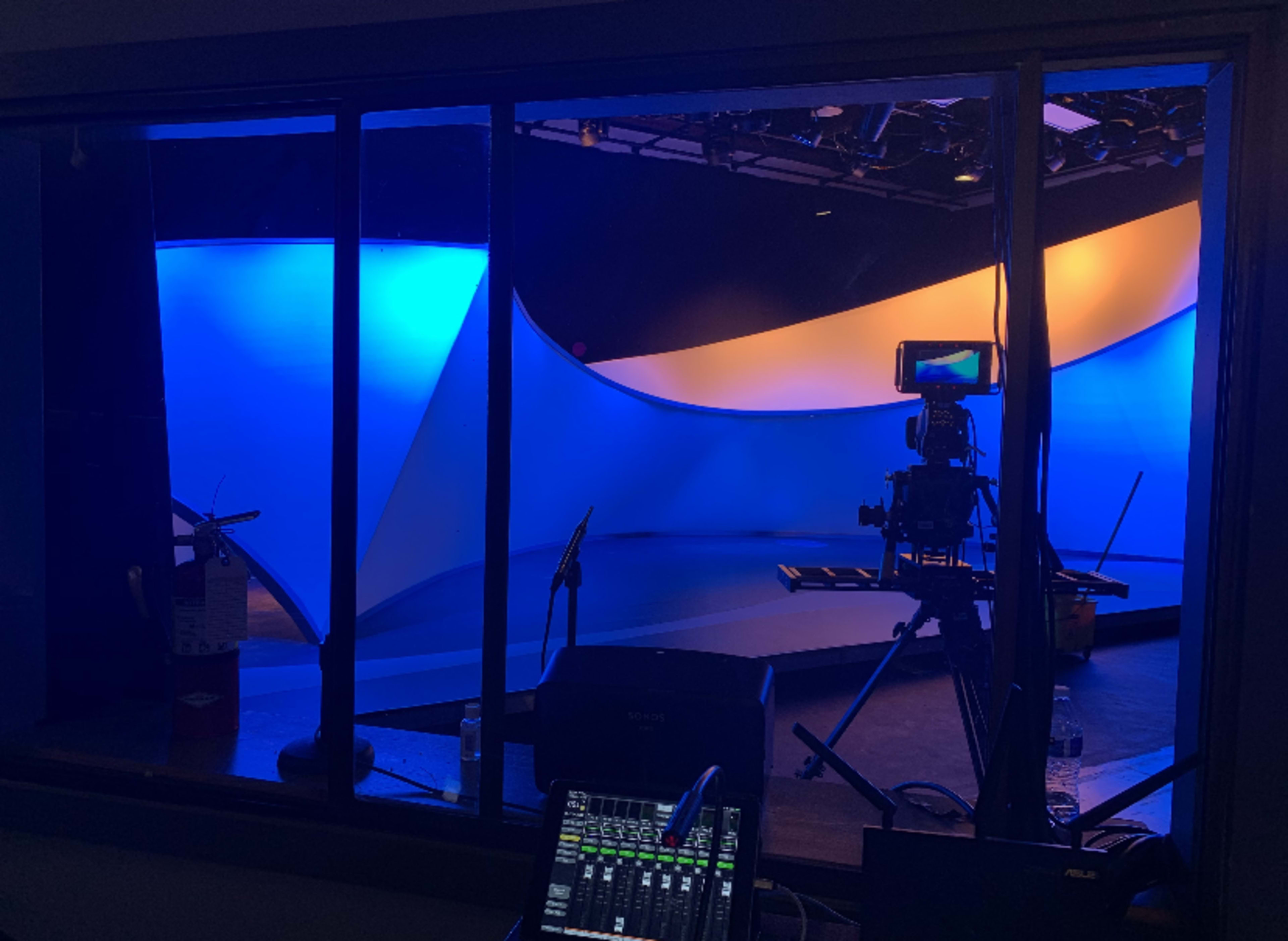 A view of a television studio set featuring curved backdrops illuminated with blue and orange lighting, along with camera equipment and a control panel in the foreground.