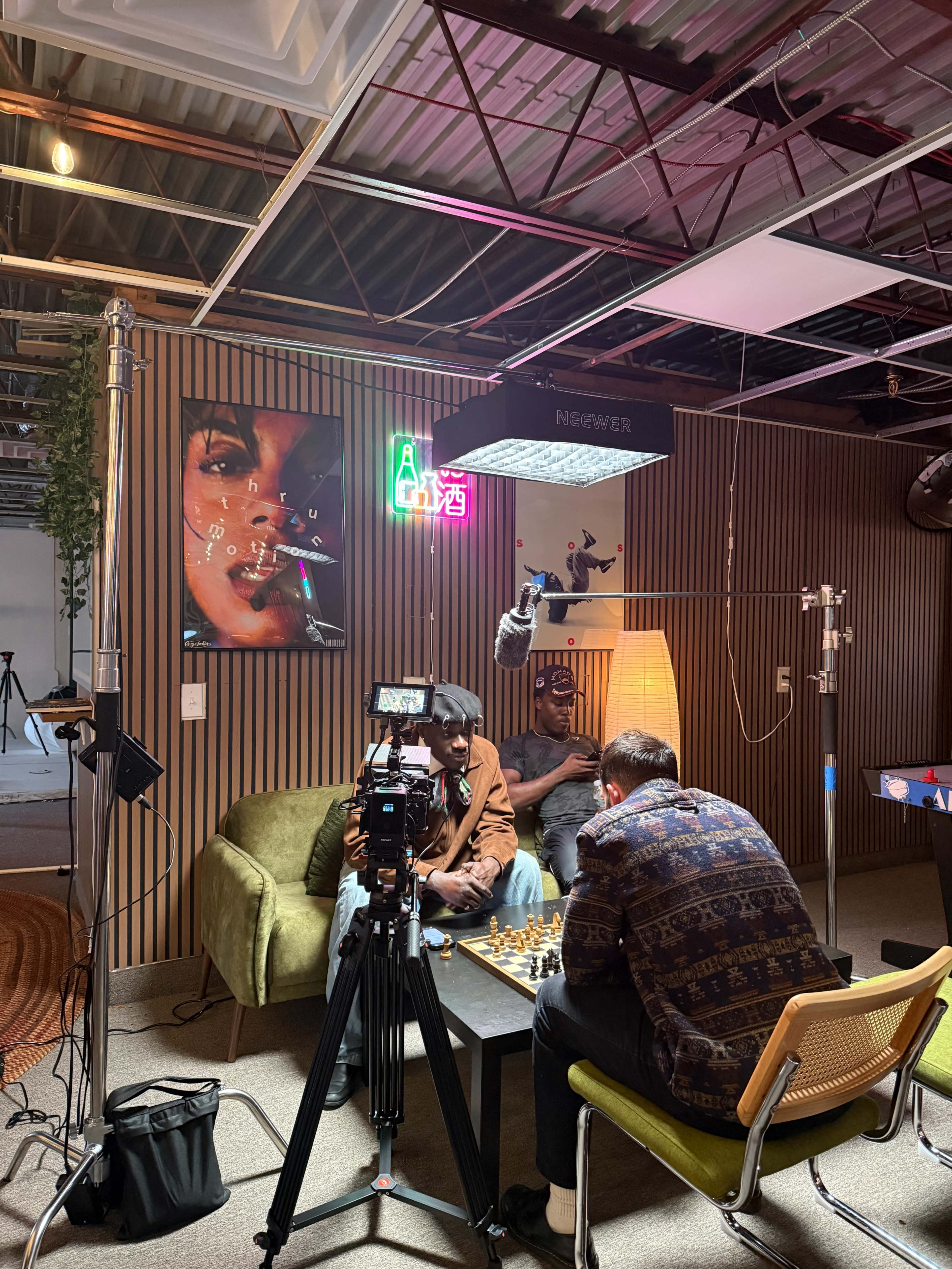 Two men play chess on a green sofa while a camera and lighting equipment are set up nearby in a studio space.