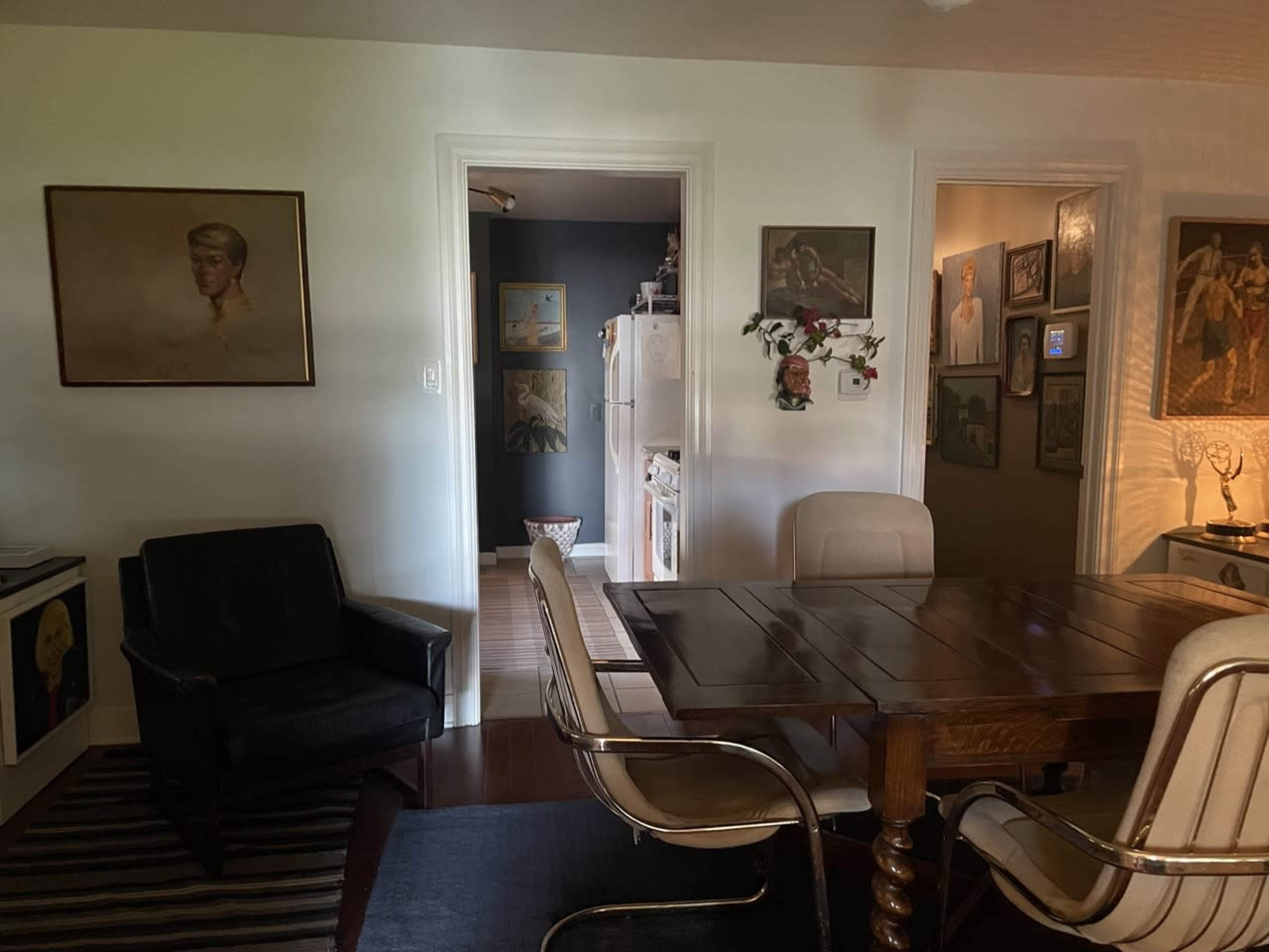The image shows a room featuring a wooden dining table surrounded by chairs, a black armchair, and two doorways leading to adjacent spaces with framed artwork on the walls.