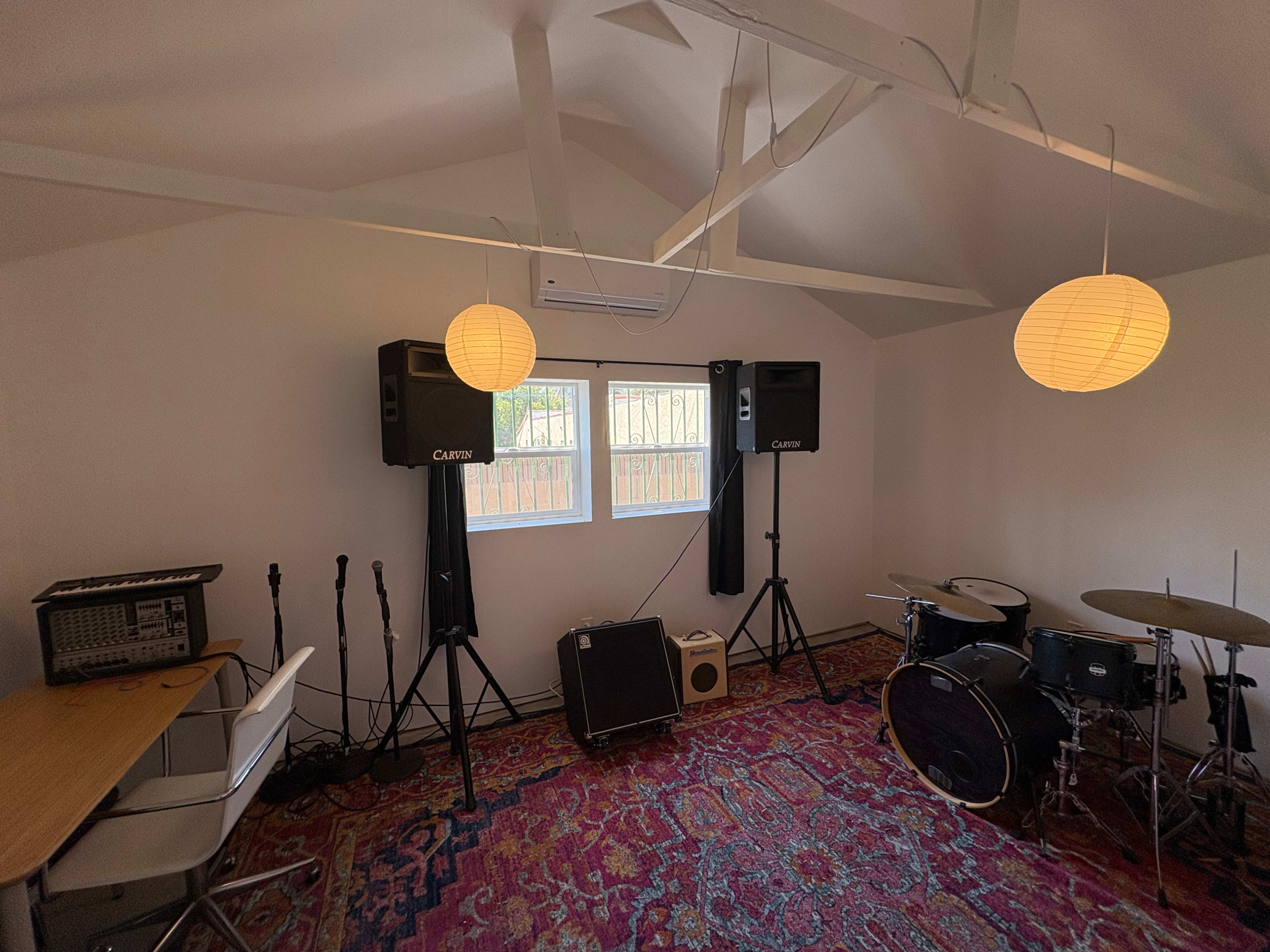 The room features a small music setup with a drum kit, speakers on stands, microphones, and a table with chairs under soft lighting.