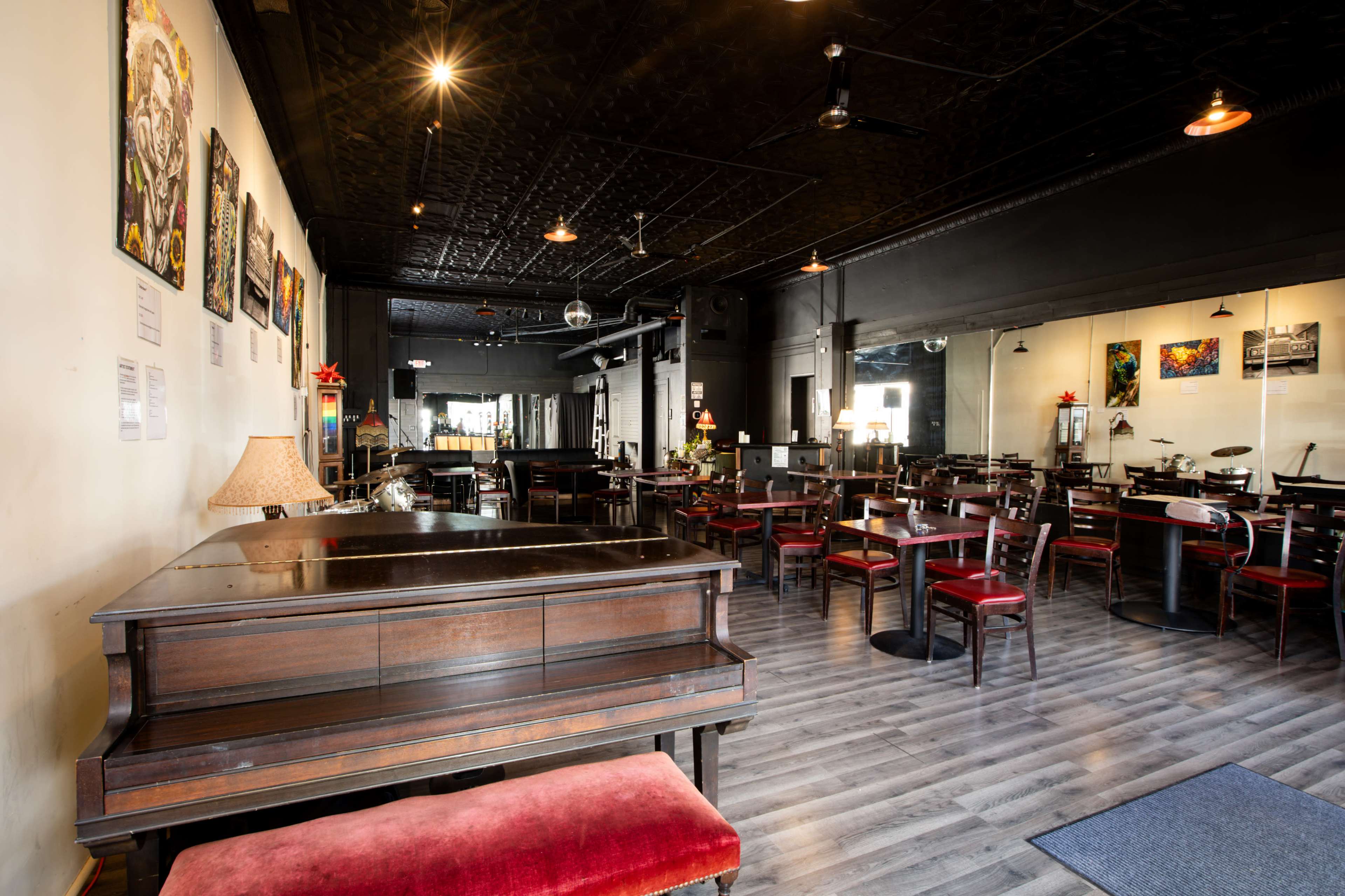 The interior of a restaurant features a grand piano, wooden tables, and chairs, with artwork adorning the walls.
