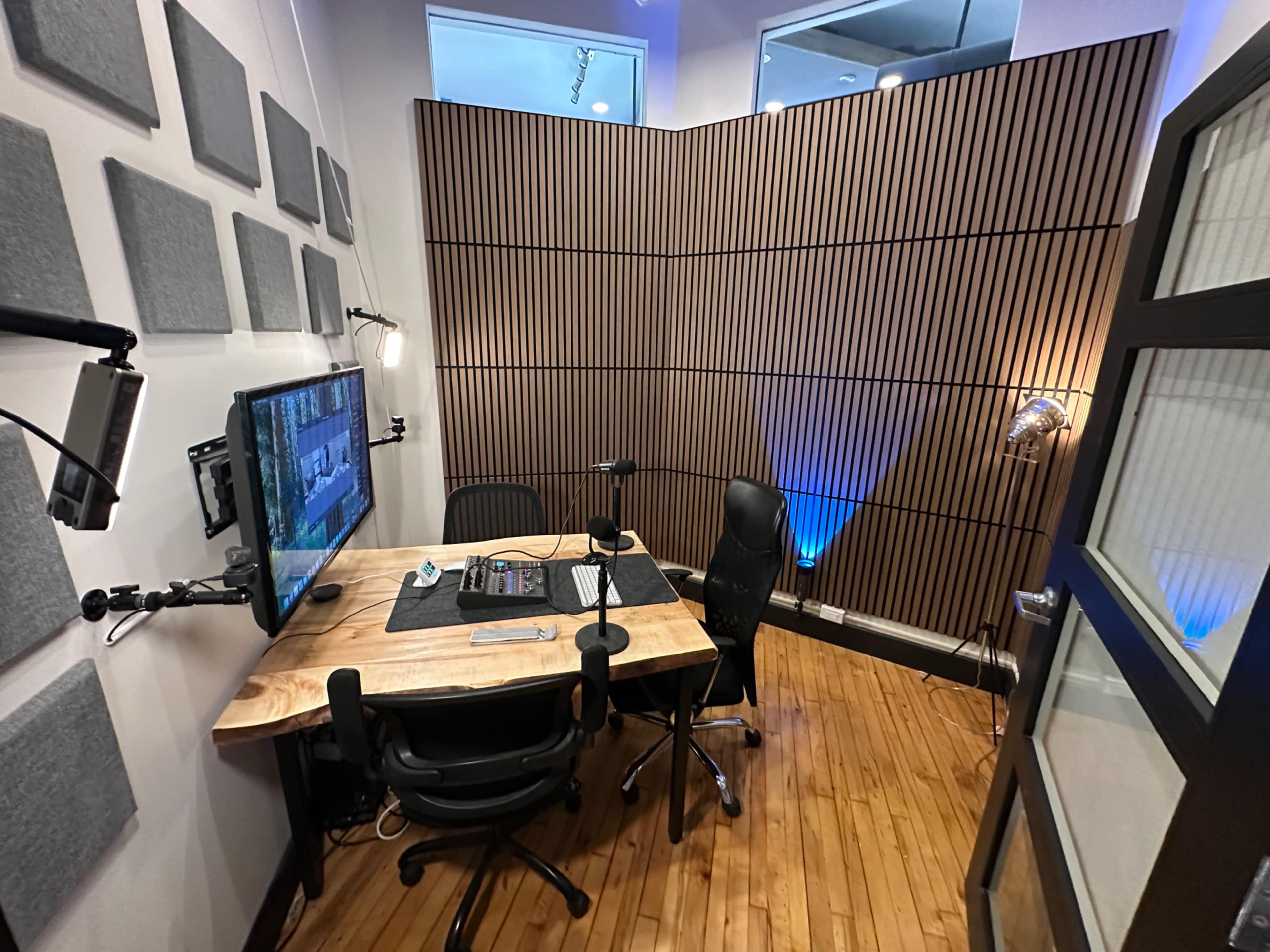The image shows a small recording studio with a wooden desk, two black chairs, a monitor on the desk, and soundproof wall panels.
