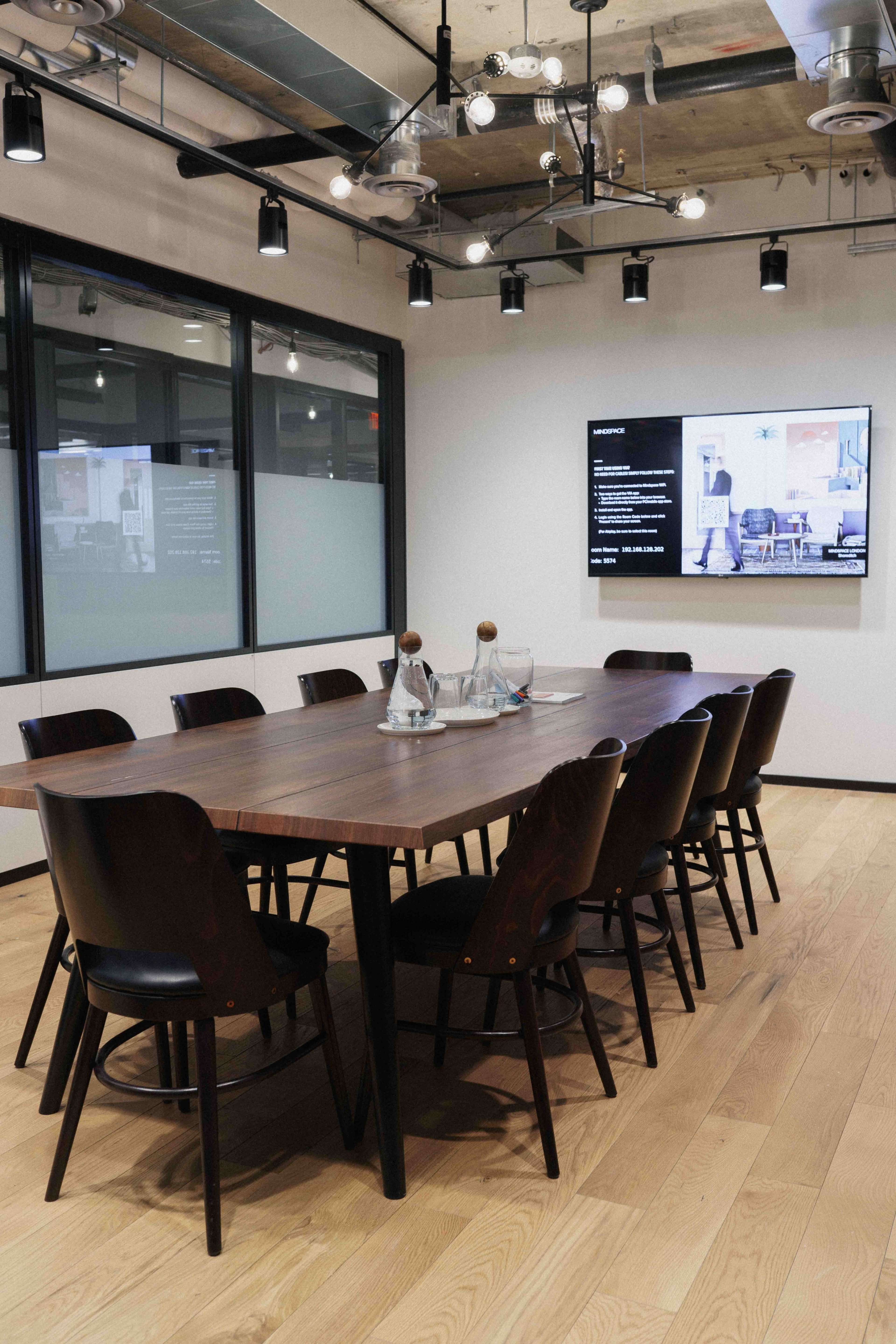 A large wooden conference table with chairs surrounds it in a modern meeting room, featuring a wall-mounted screen displaying information.