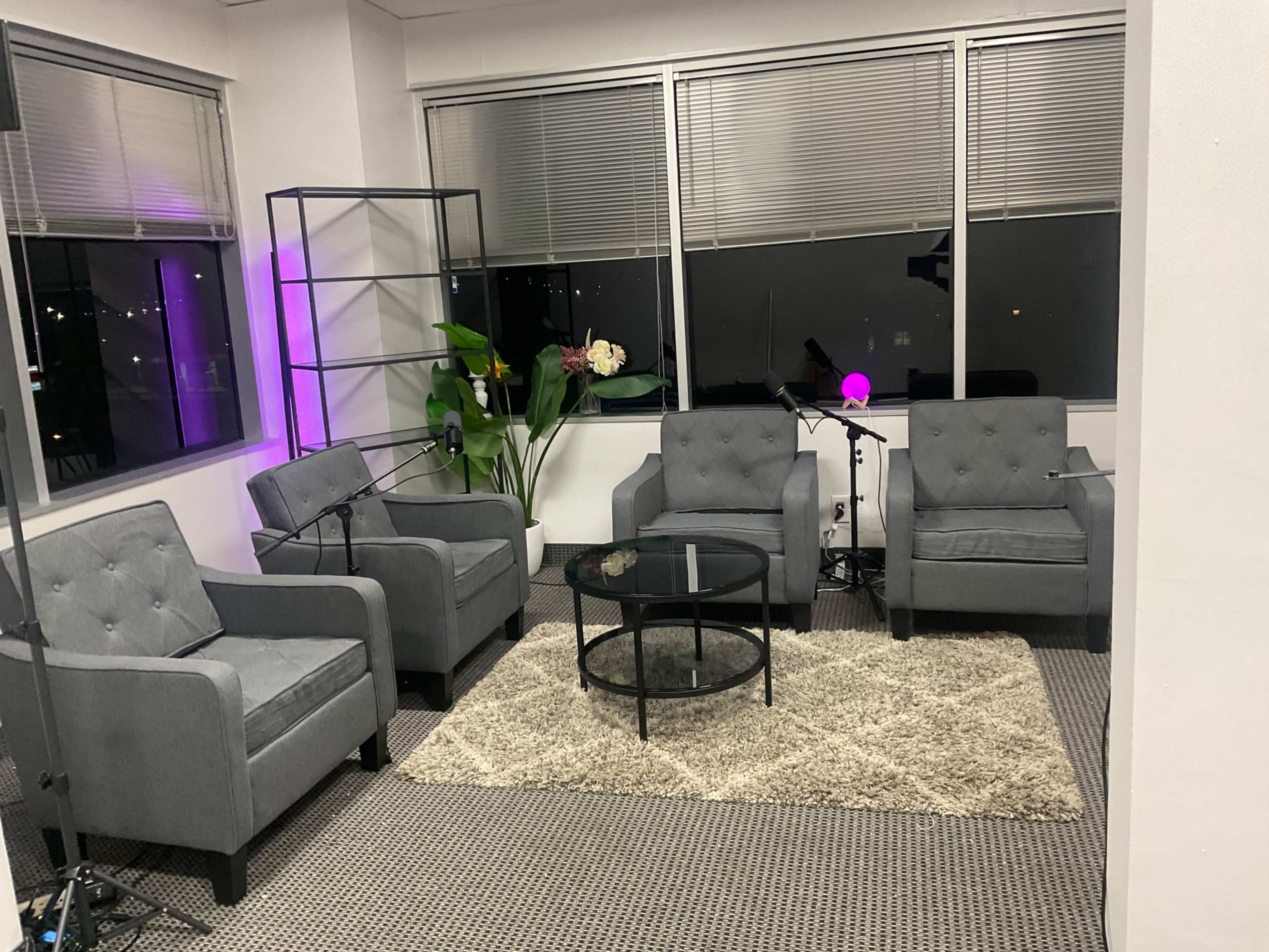 The image shows a modern seating area with four gray chairs arranged around a circular glass coffee table, placed on a light-colored rug, with a plant and purple ambient lighting in the background.