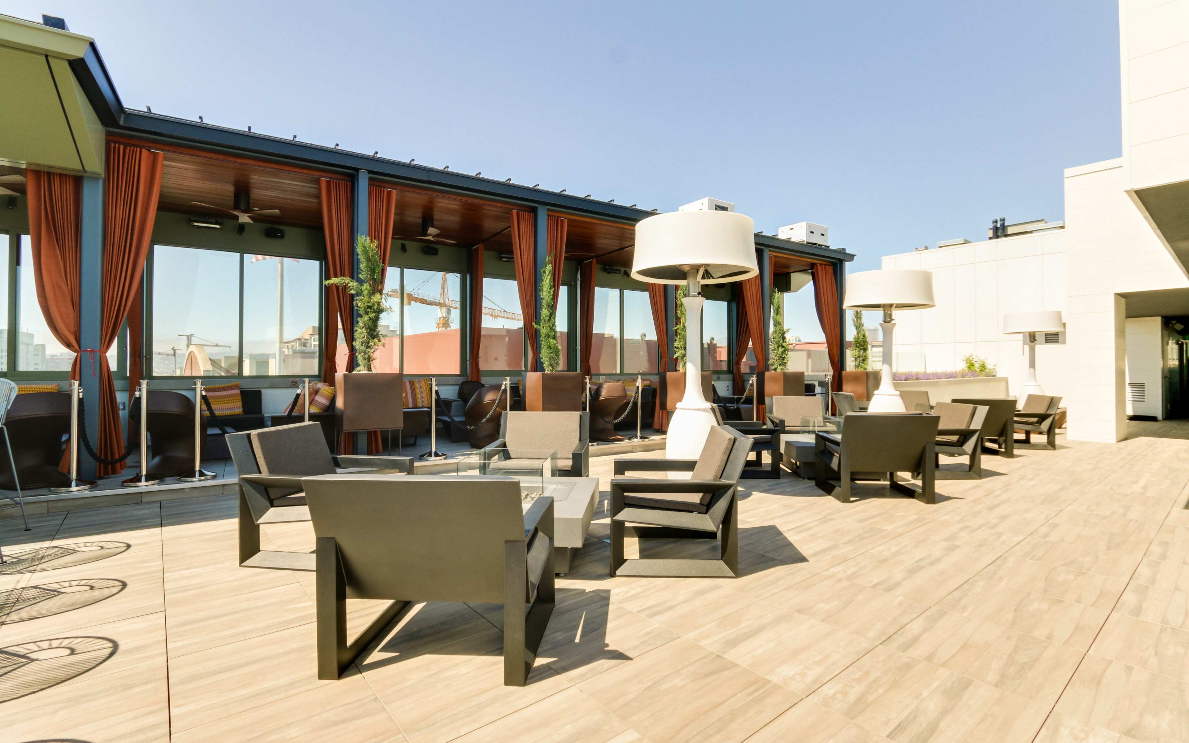 A rooftop lounge area with several chairs and tables, surrounded by large windows and draped curtains.
