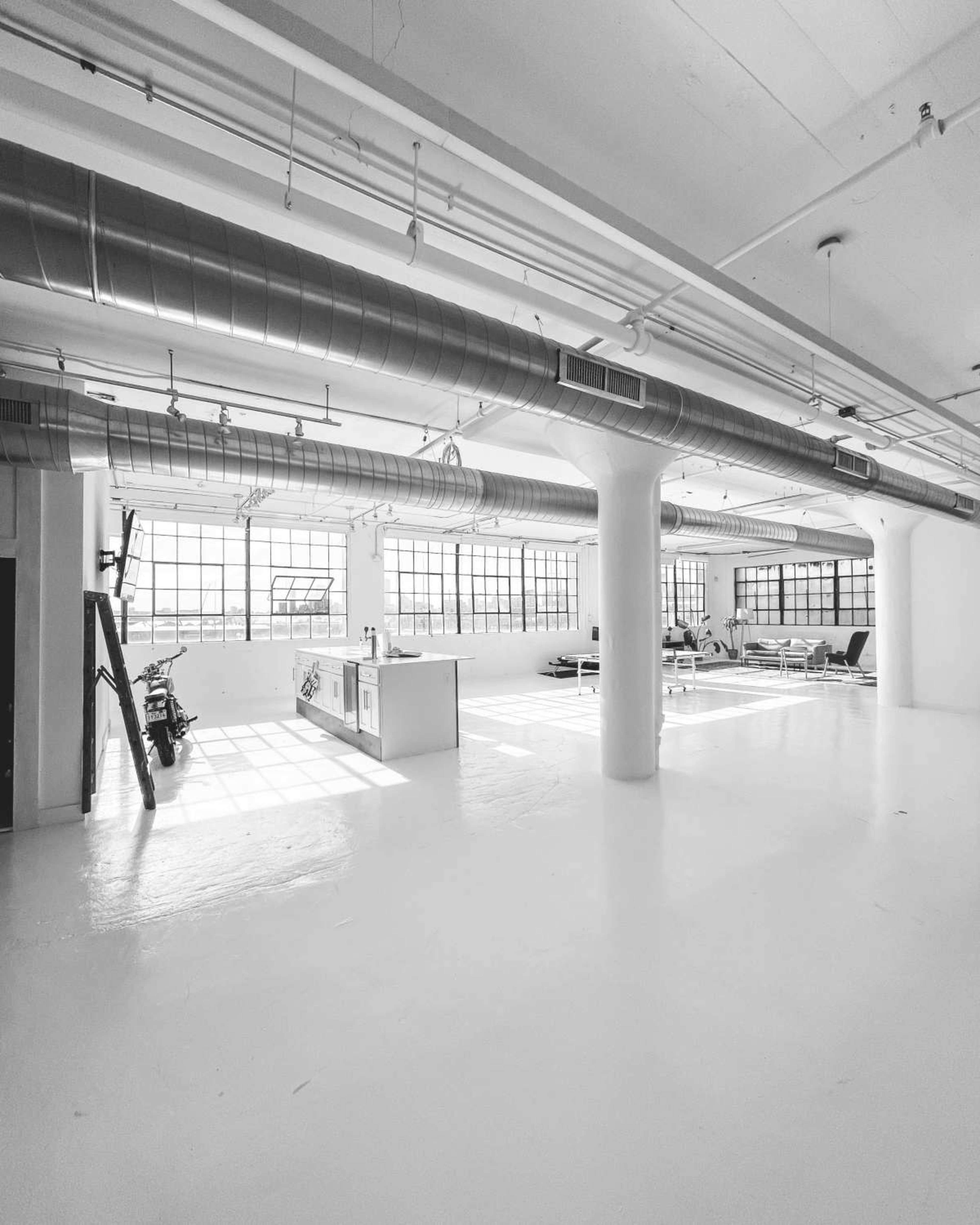 The image shows a spacious, industrial-style room with large windows, exposed ducts, and a kitchen area on one side.