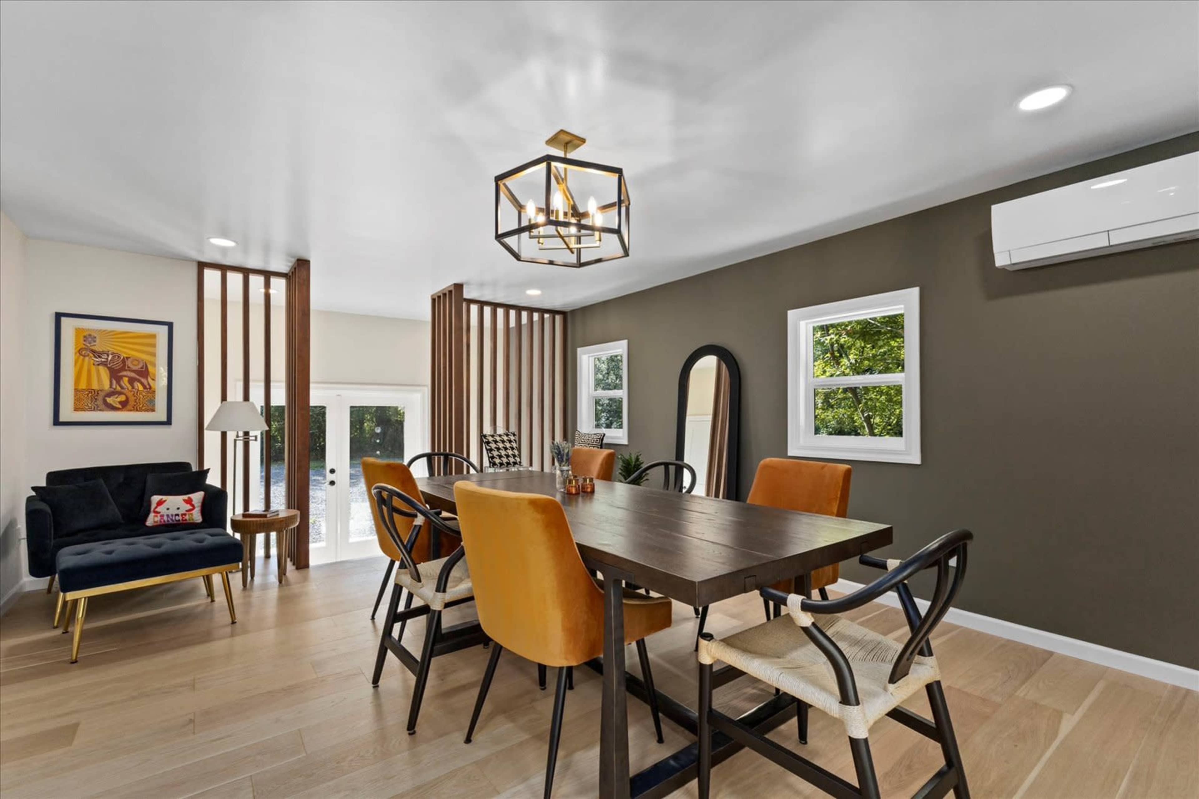 The image shows a modern dining area featuring a wooden table surrounded by orange and black chairs, with large windows and decorative wall elements.