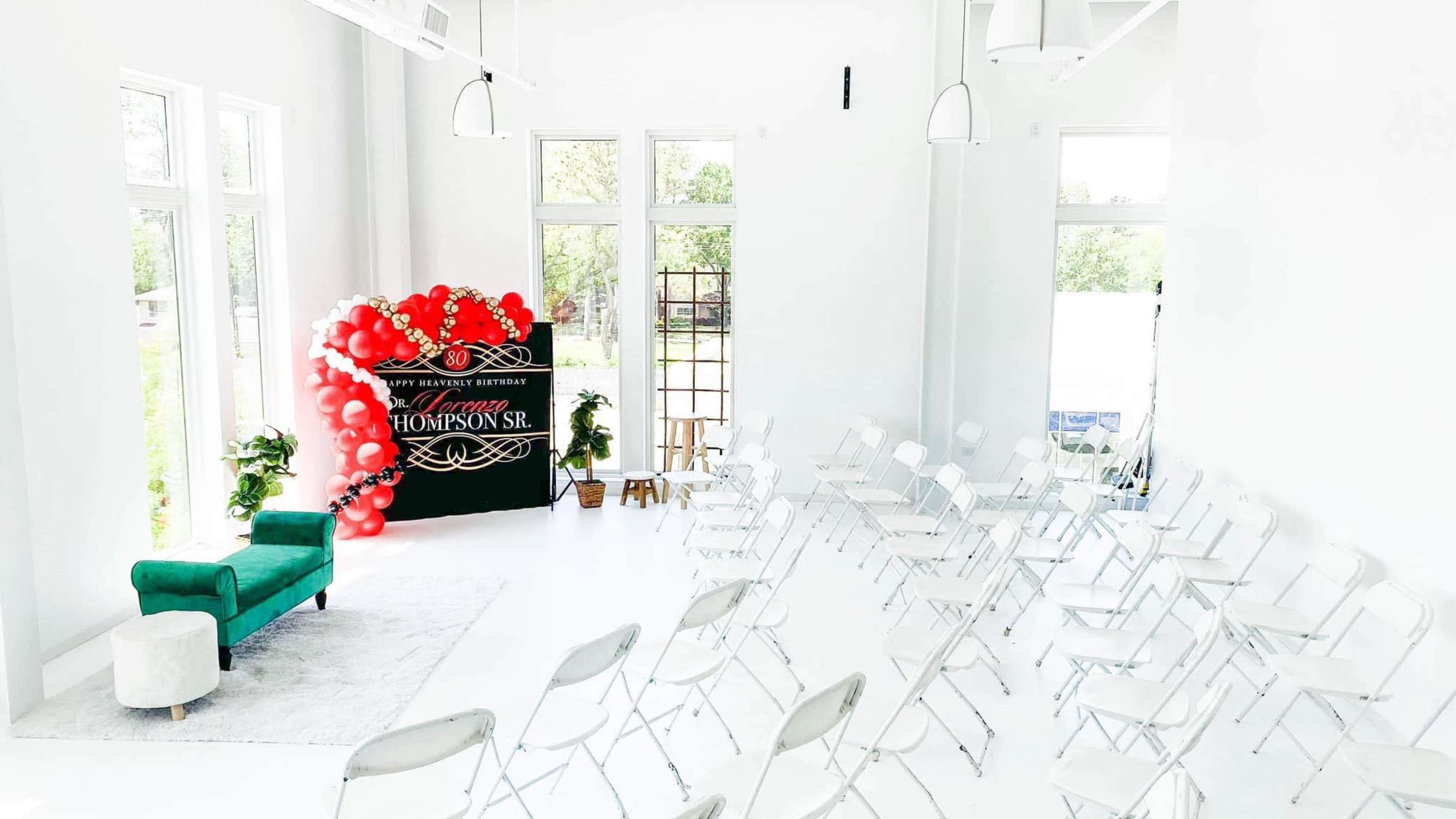 A spacious event room with white walls, a row of silver folding chairs arranged for seating, and a decorative backdrop featuring red balloons and the name "Thompson Sr." over a green sofa.
