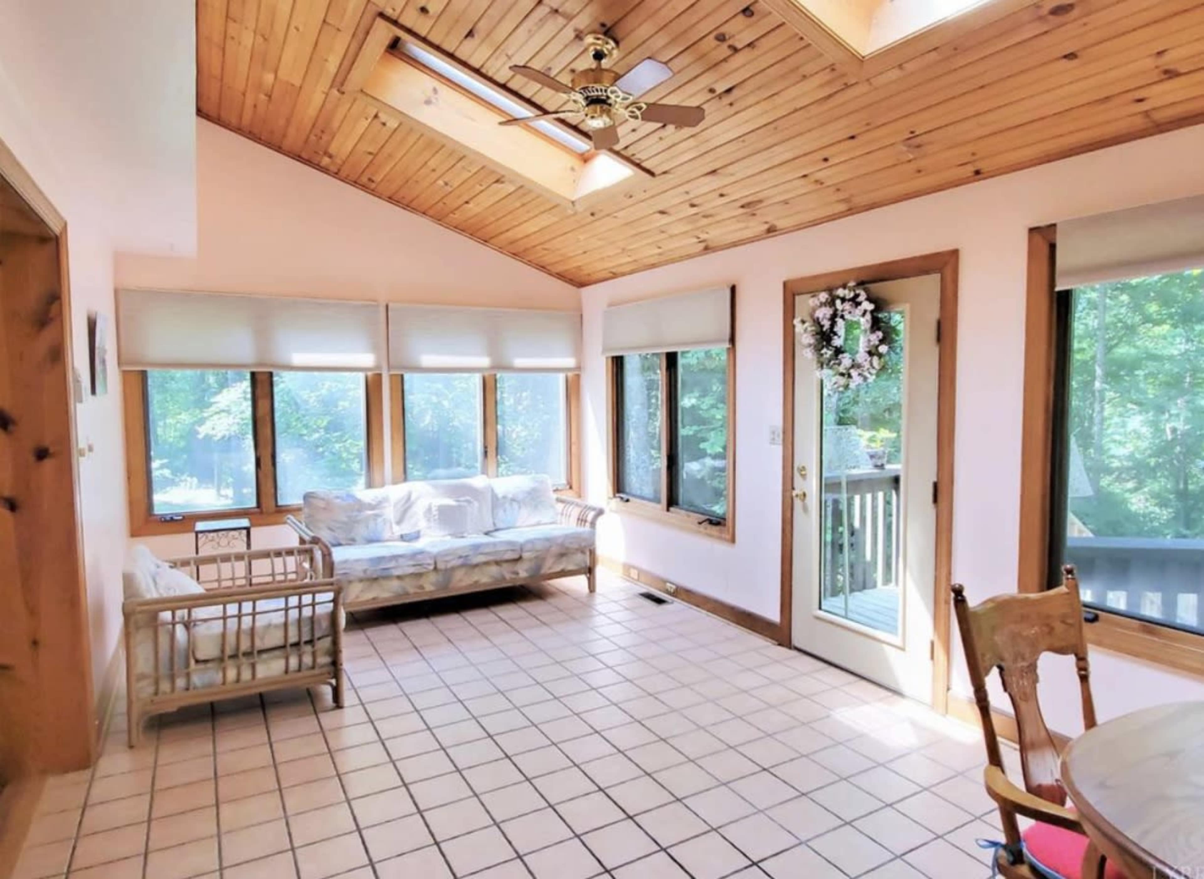 A sunlit room features a wooden ceiling, large windows, a couch, and access to a deck through a door.