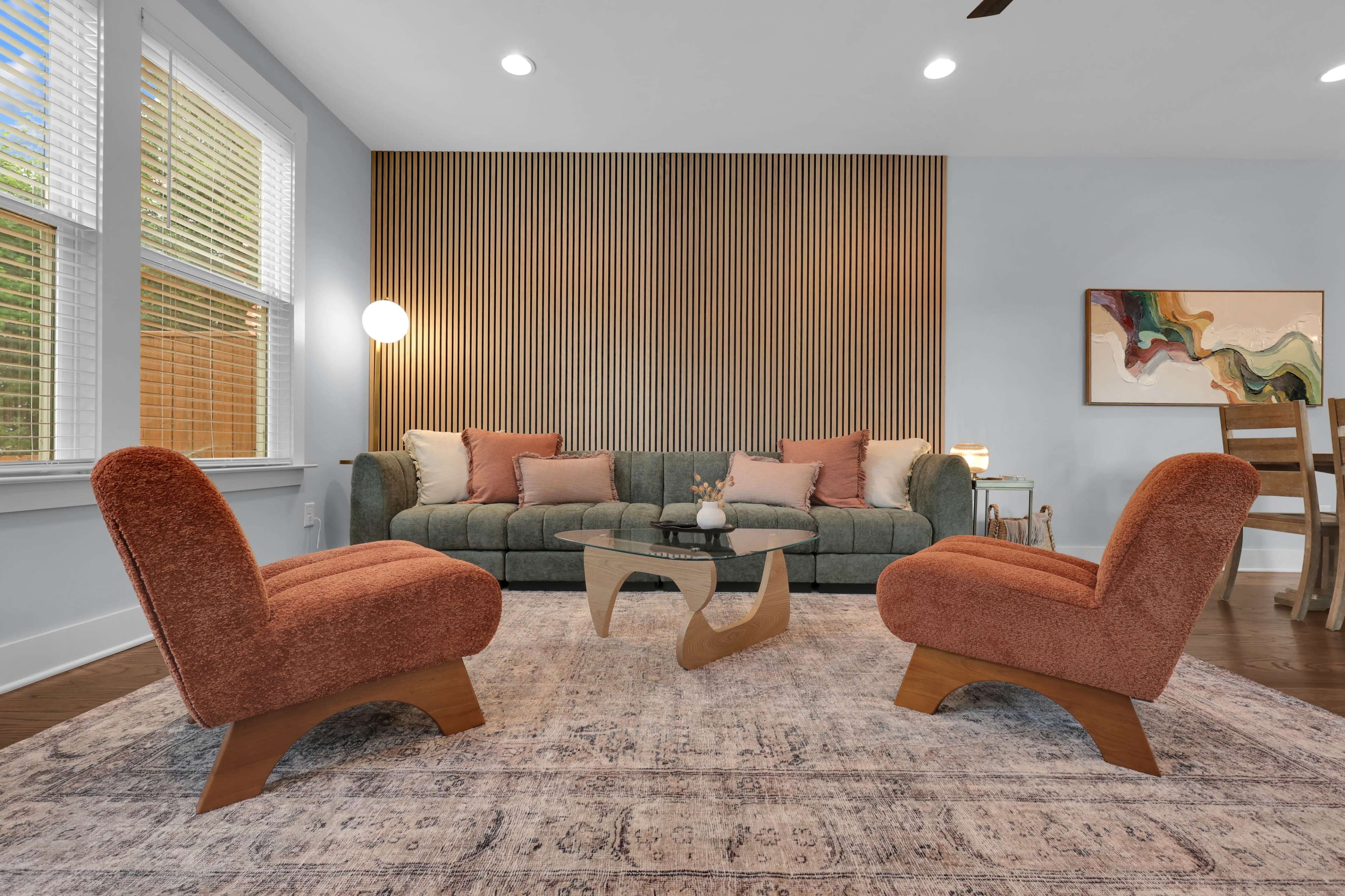 The scene features a cozy living room with a green sofa, two orange lounge chairs, and a glass coffee table on a patterned area rug, complemented by wooden accents and natural light from the windows.