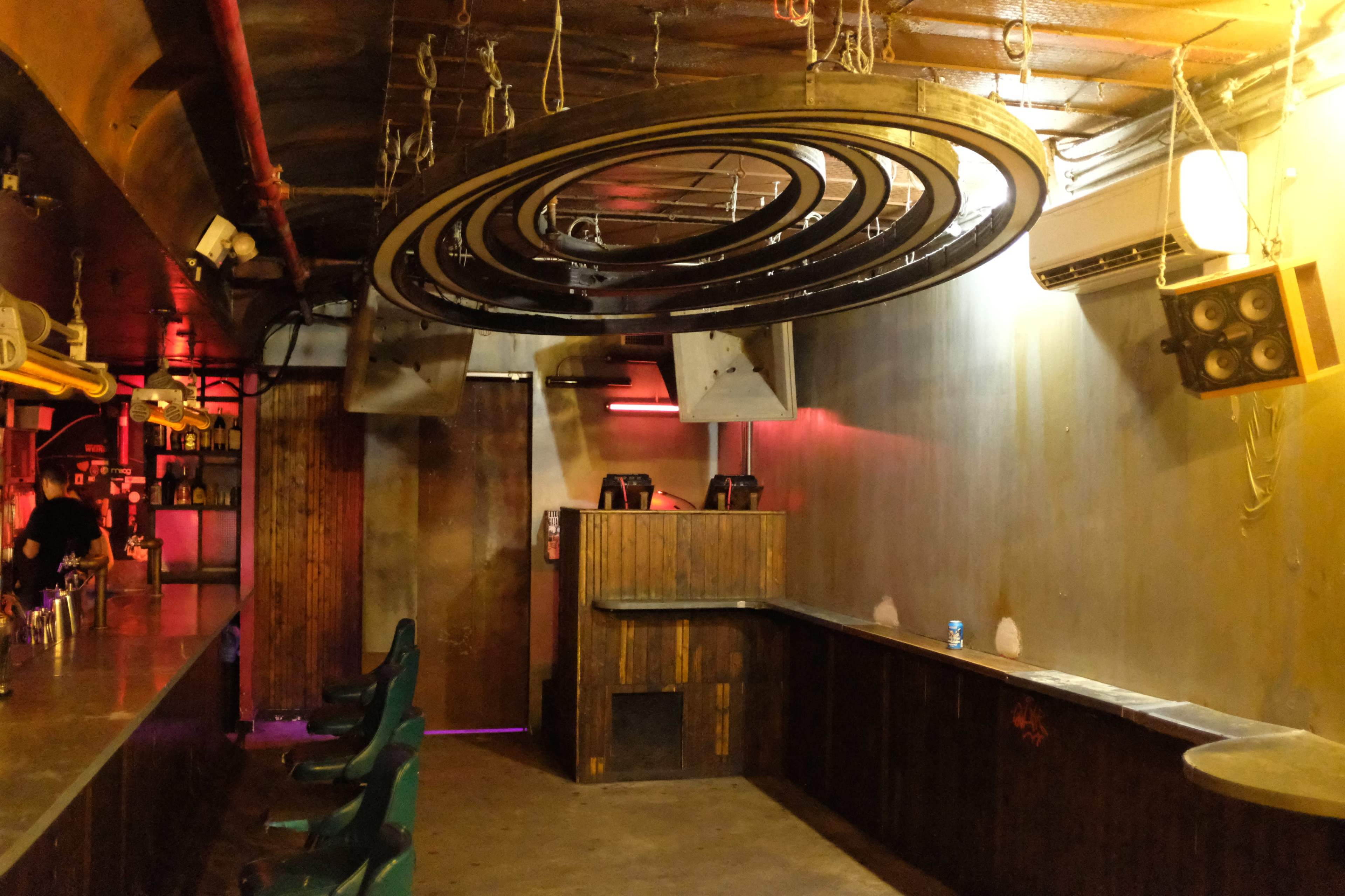 The image shows a dimly lit bar interior featuring a circular hanging light fixture, a wooden counter, and a few green chairs.