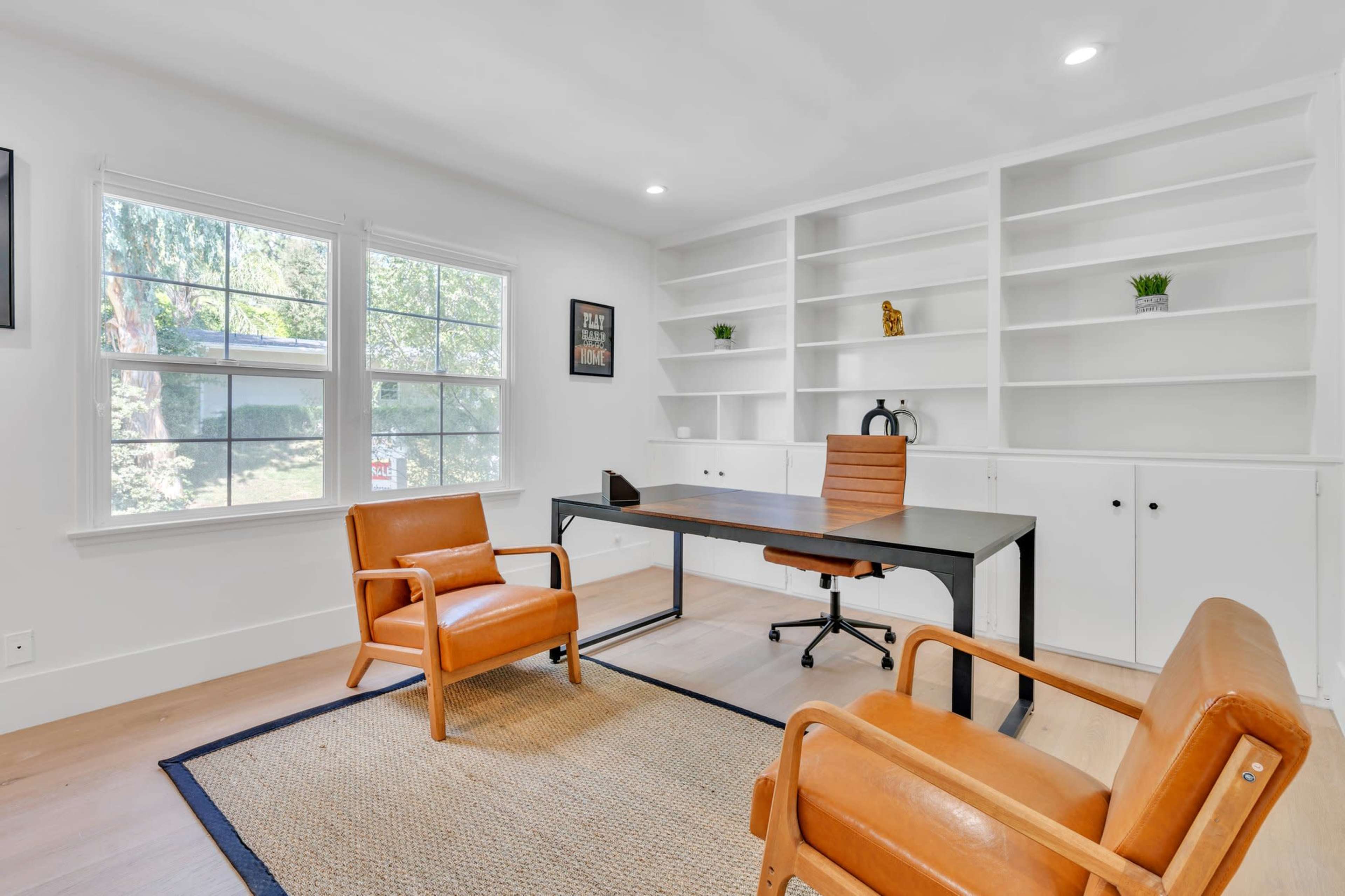 The image shows a bright, minimalist office space featuring a wooden desk, two orange leather chairs, and built-in shelves against the wall.