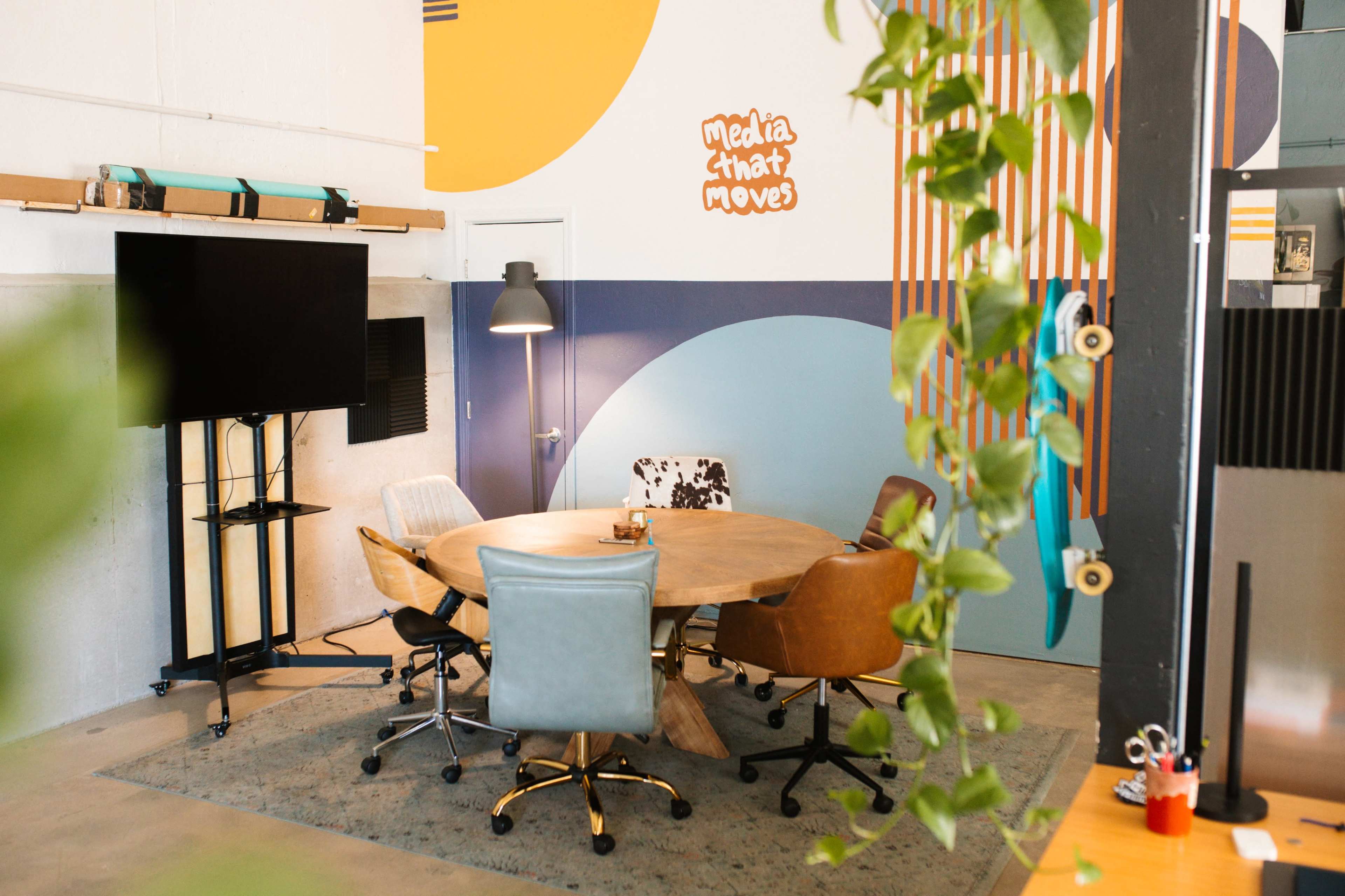 The image shows a modern conference room with a round wooden table surrounded by various chairs, a television on a stand, and colorful wall graphics.