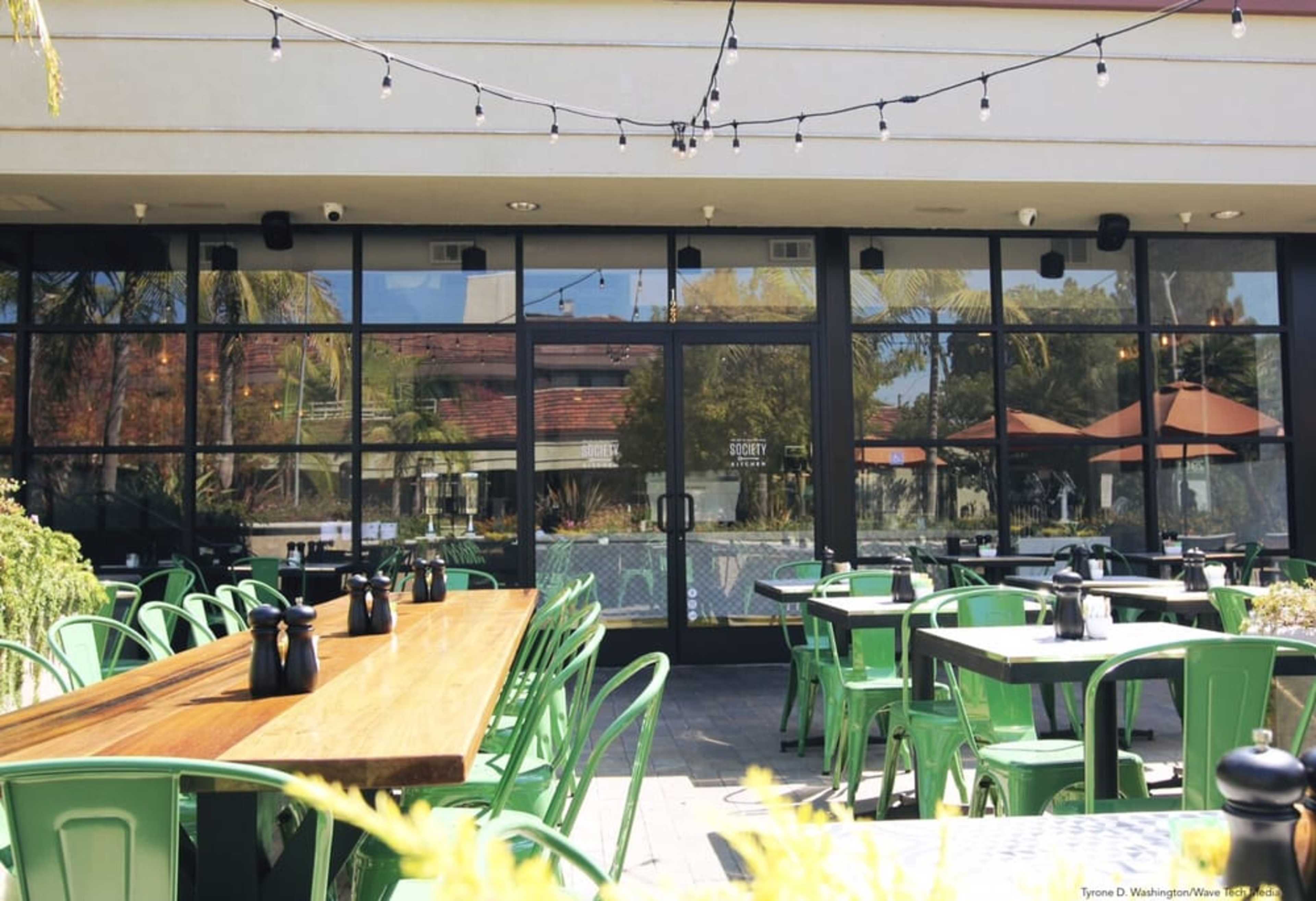 A patio area with green metal chairs and wooden tables, overlooking large glass doors that reflect the surroundings.
