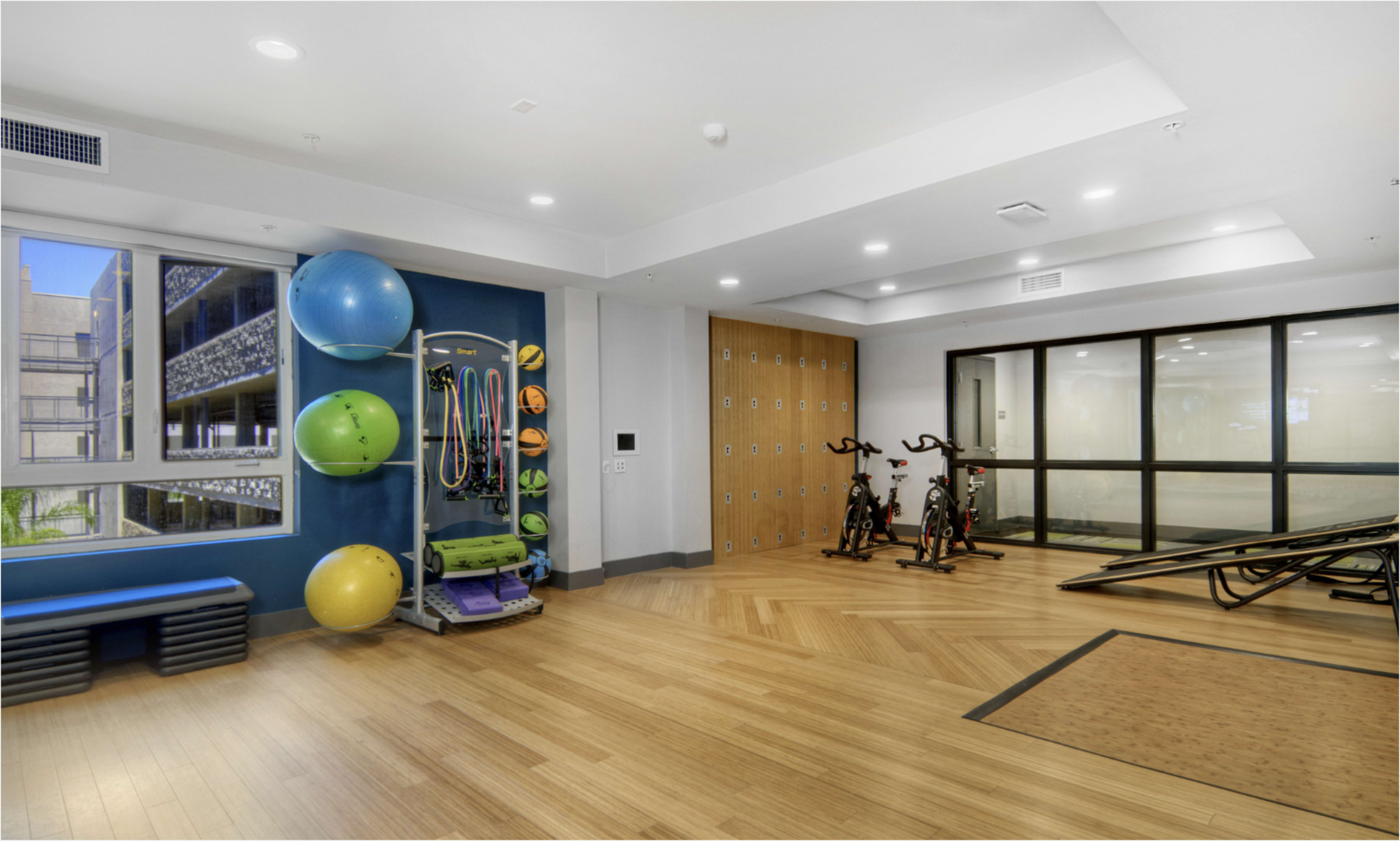 The image showcases a fitness room featuring exercise equipment, exercise balls, and lockers, with large windows providing natural light.