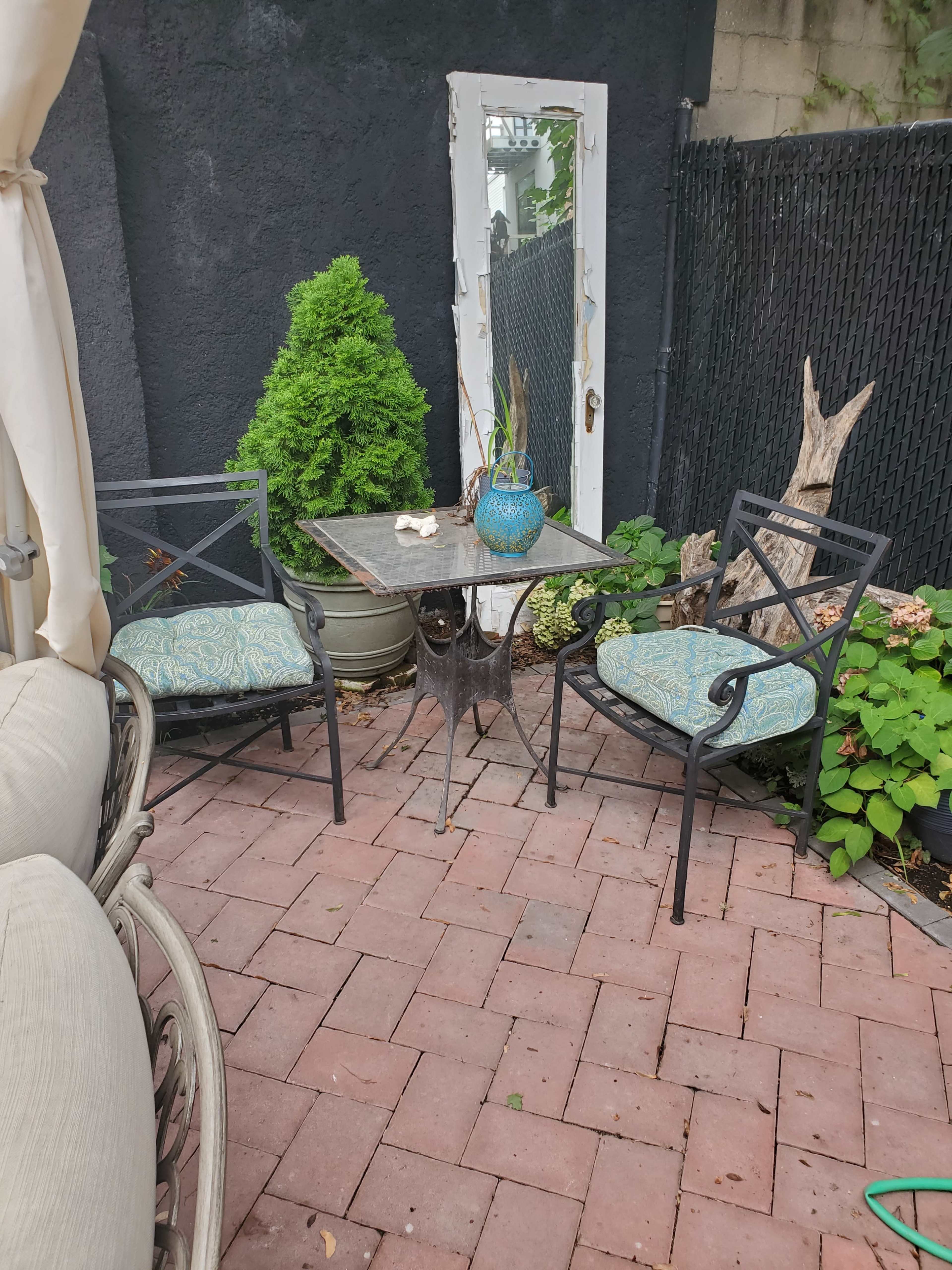 A small outdoor seating area features two metal chairs and a table set on a brick patio, with a mirror and potted plants in the background.