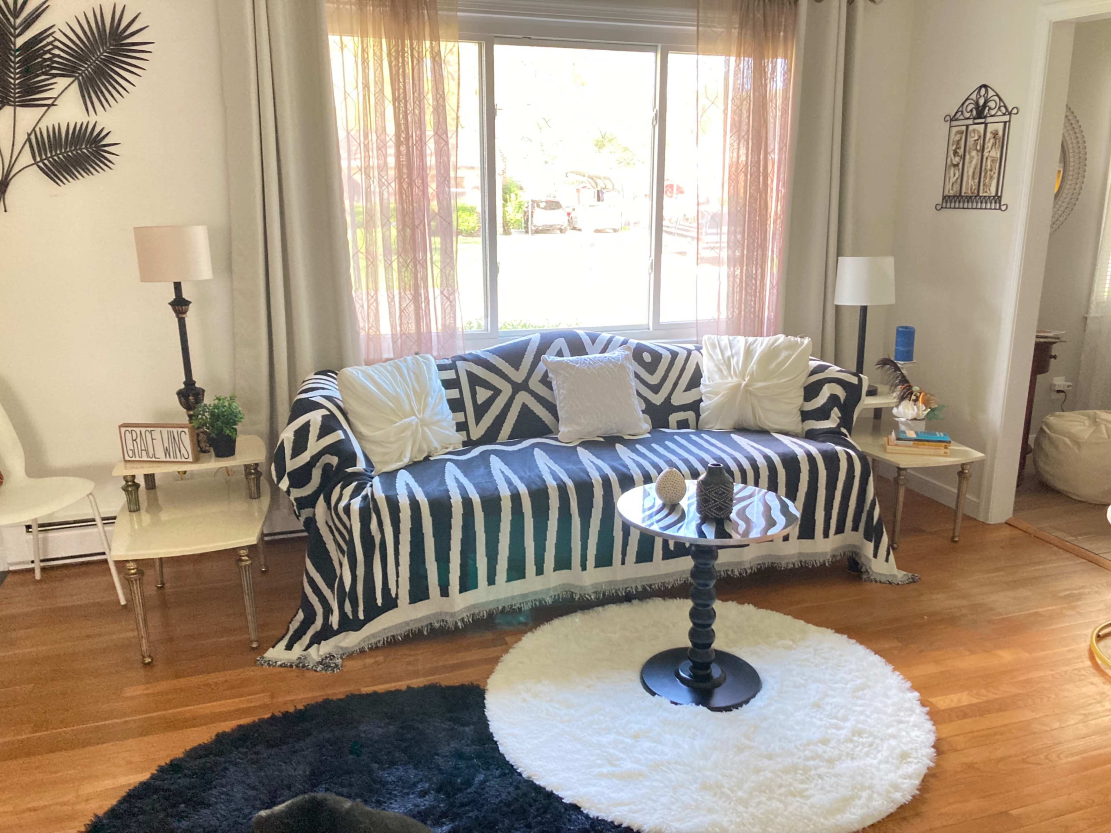 A living room features a patterned couch with decorative pillows, a round table with a black base, and a fluffy white rug under a large window.