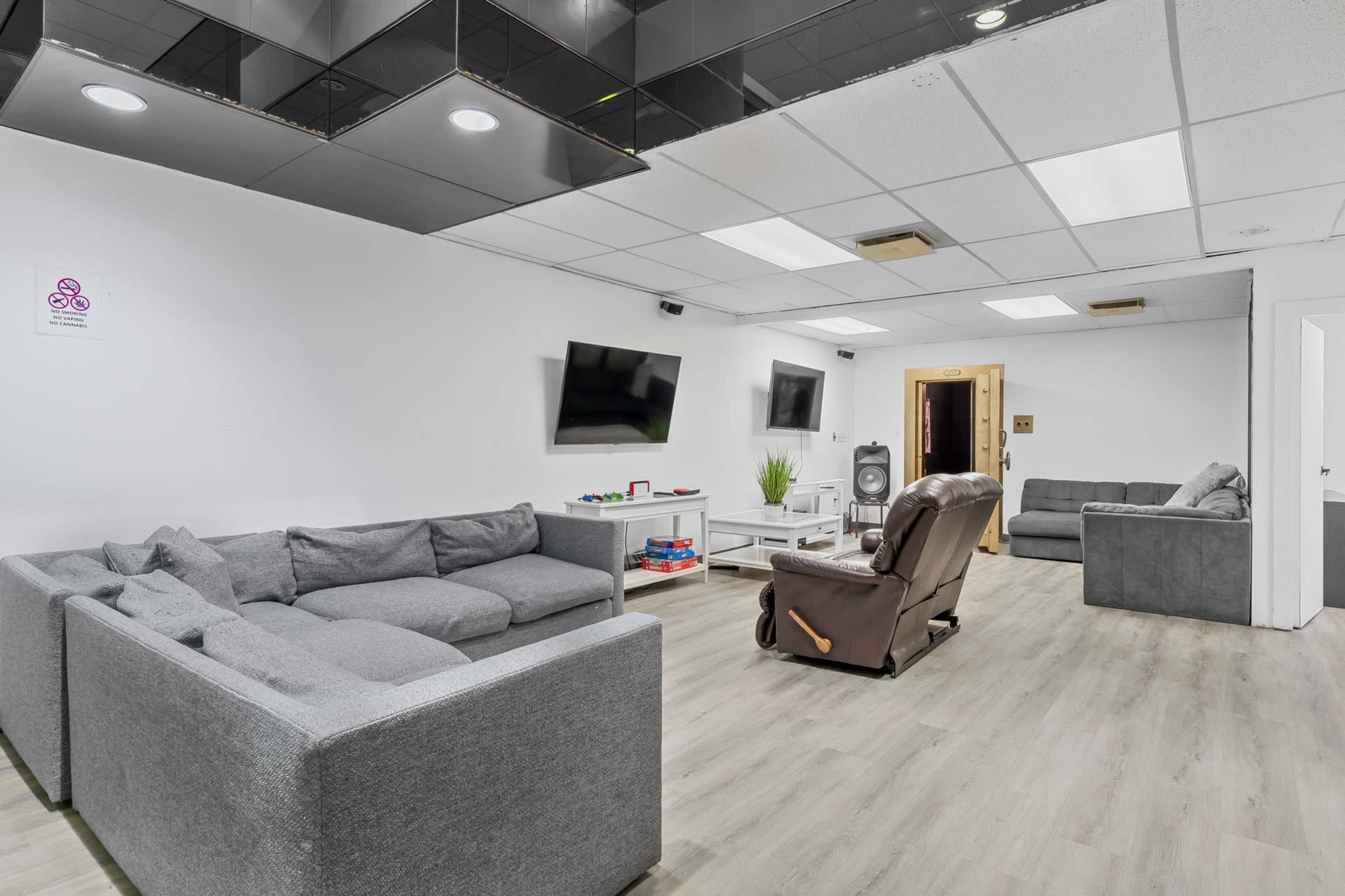 A spacious lounge area features gray sectional sofas, a recliner, two wall-mounted TVs, and light wood flooring.