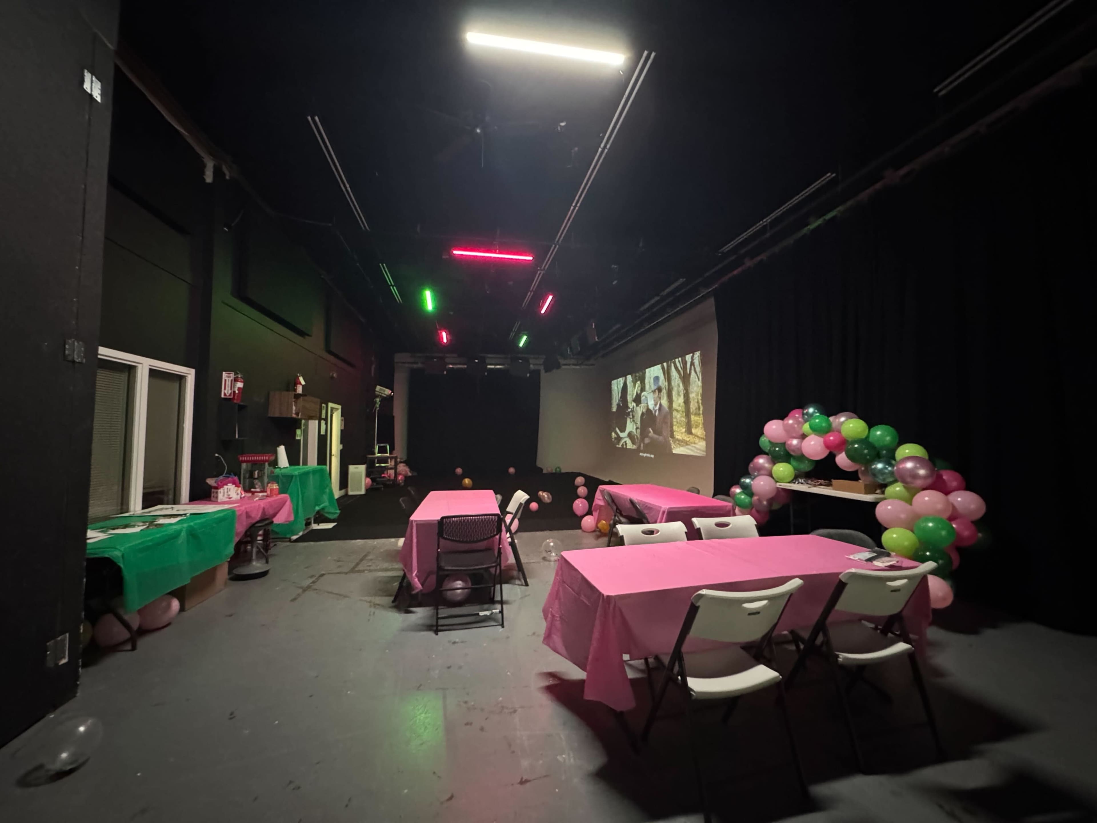The image shows an event space with tables covered in pink and green tablecloths, a balloon arch, and a large screen displaying a video in a dimly lit environment.