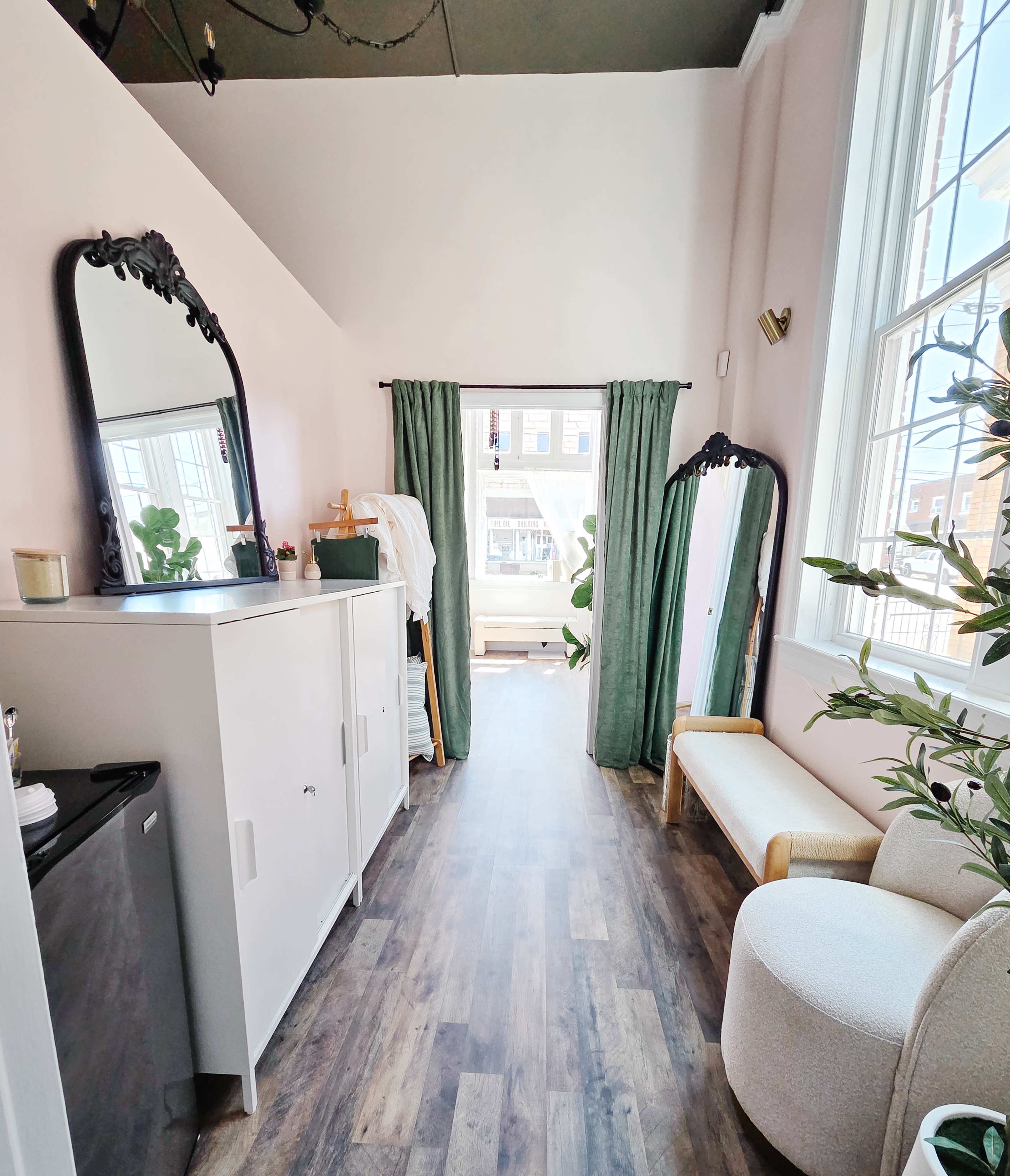 The image shows a bright interior hallway with a mirrored cabinet, curtains, and a seating area, leading to a window with natural light.