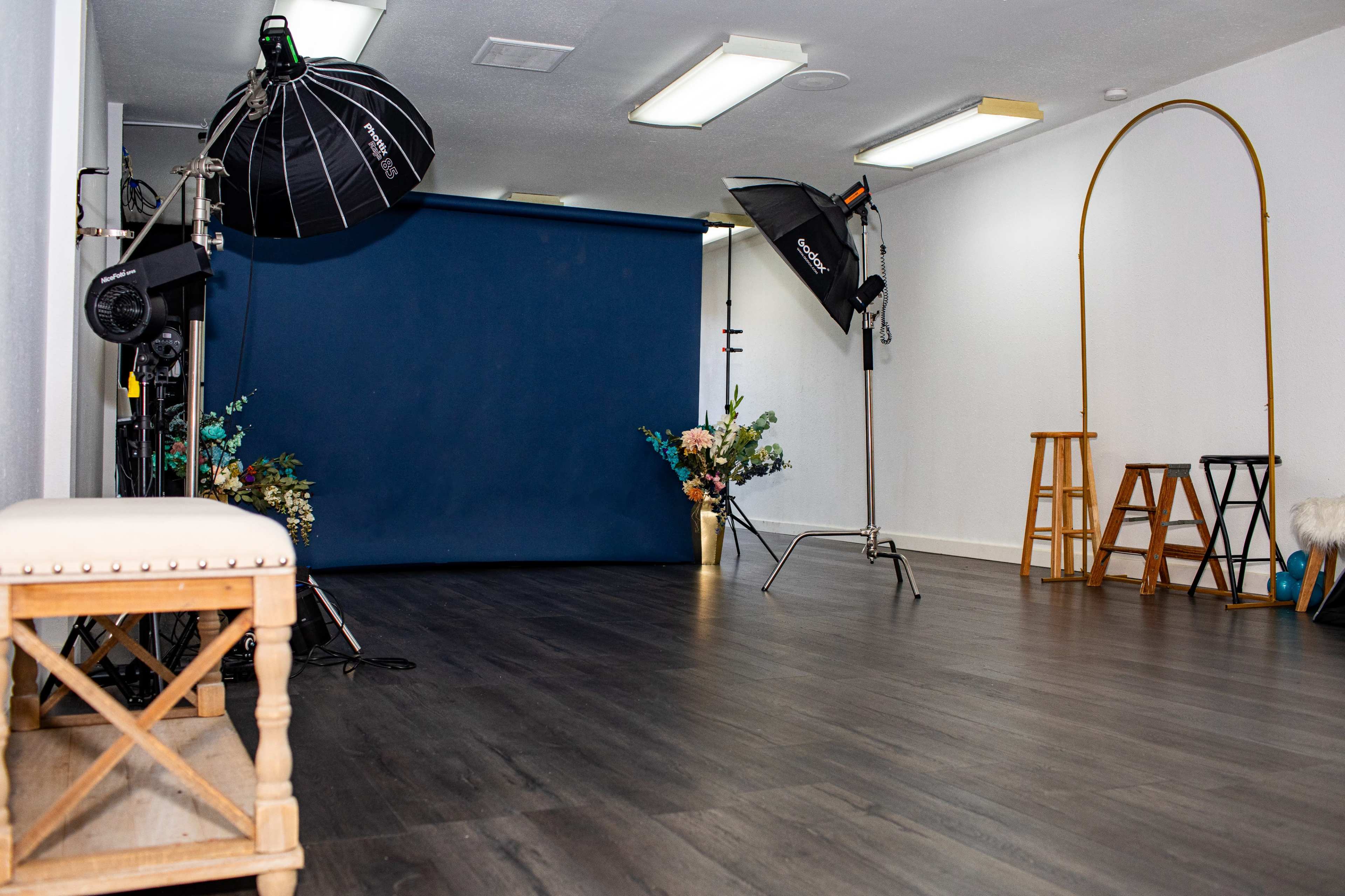 The image shows a photography studio featuring a blue backdrop, several lighting equipment setups, and minimal decor.