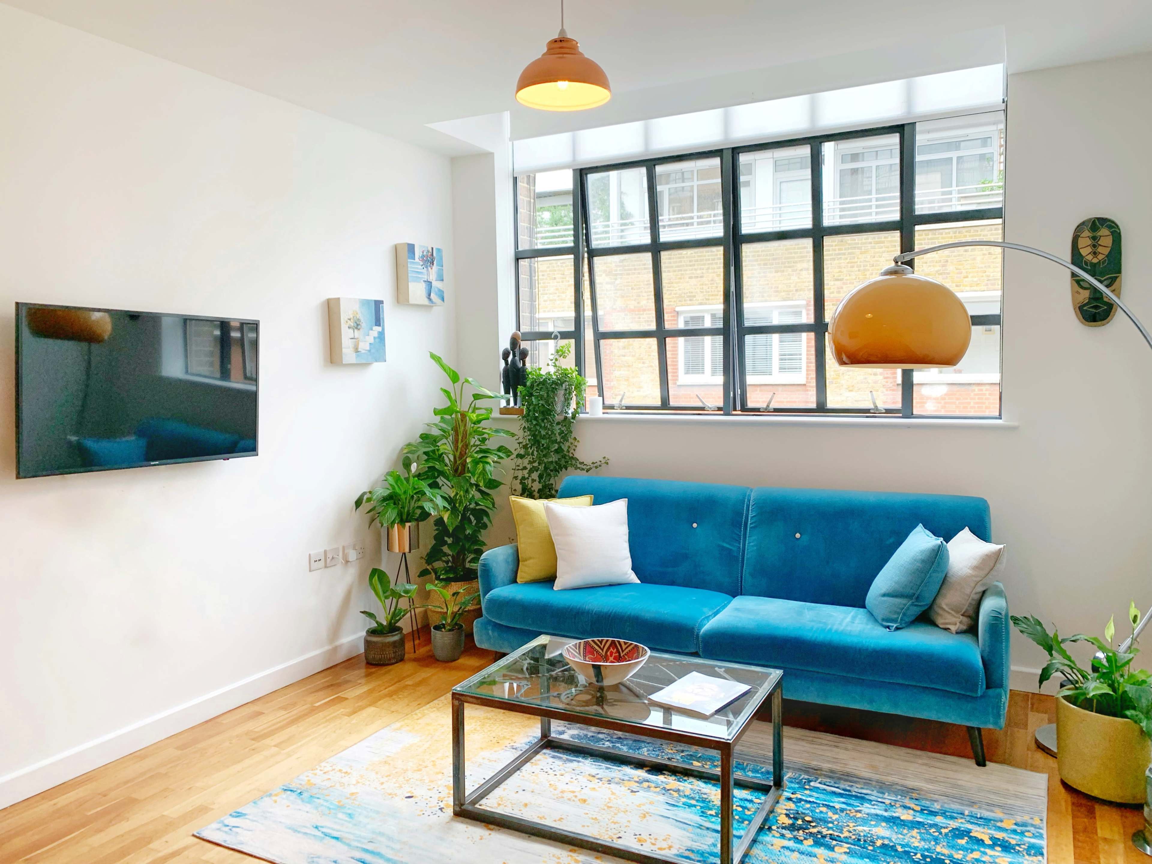 A bright living room features a blue sofa, a glass coffee table, a television mounted on the wall, and various indoor plants.