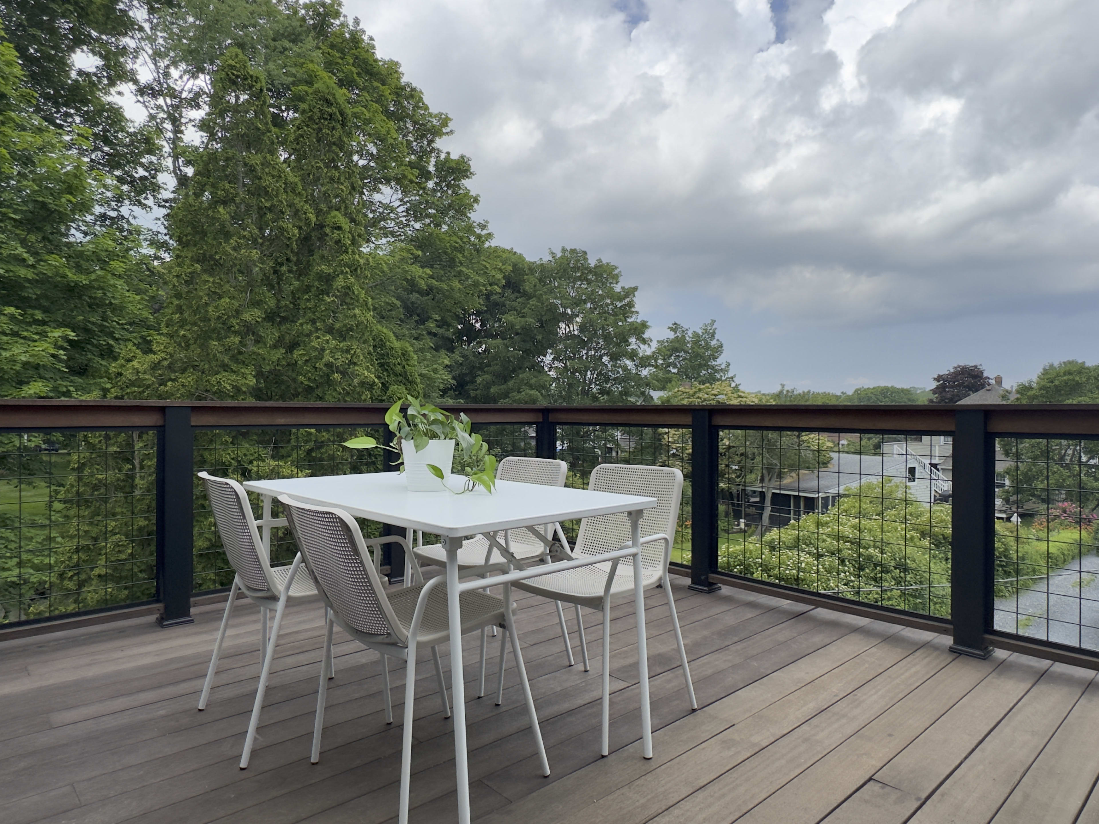 A white table and six chairs are set on a wooden deck surrounded by greenery under a cloudy sky.