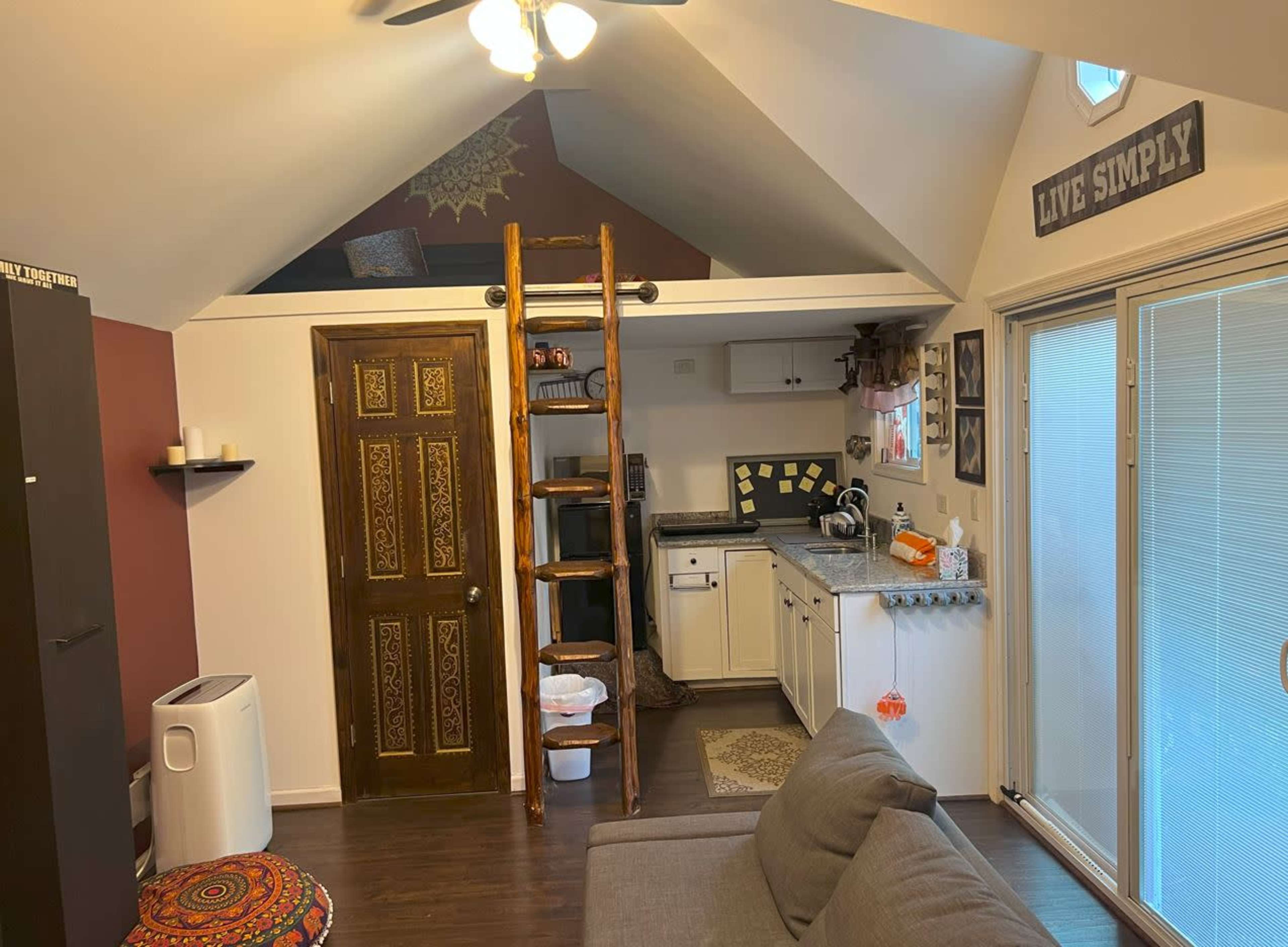 A small, cozy living area with a lofted sleeping space accessed by a wooden ladder, a kitchenette, and sliding glass doors leading outside.