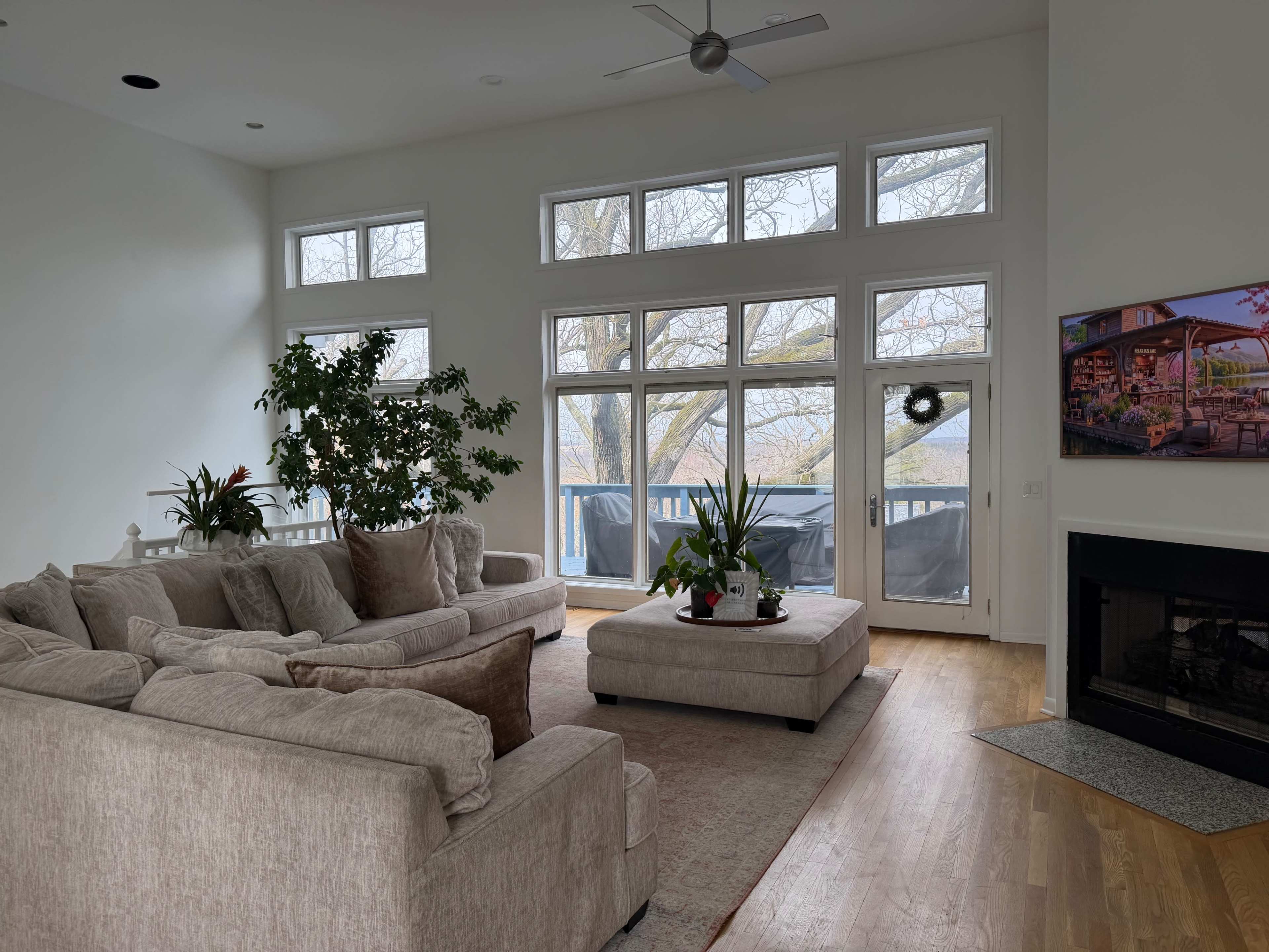 A spacious living room features a large sectional sofa, a coffee table with plants, and a wall-mounted TV, all with large windows providing views of a deck and trees outside.