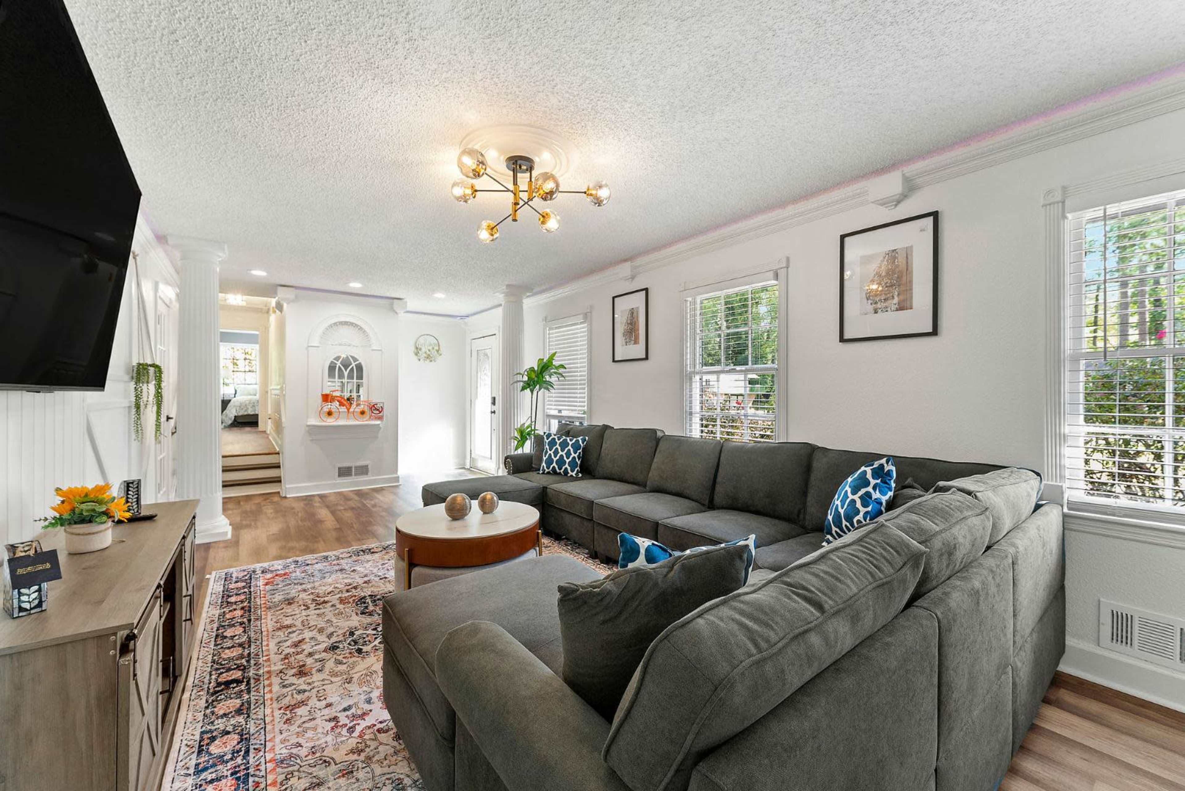 A modern living room features a large gray sectional sofa, a round coffee table, and decorative art on the walls, with a bright ambiance provided by natural light from the windows.