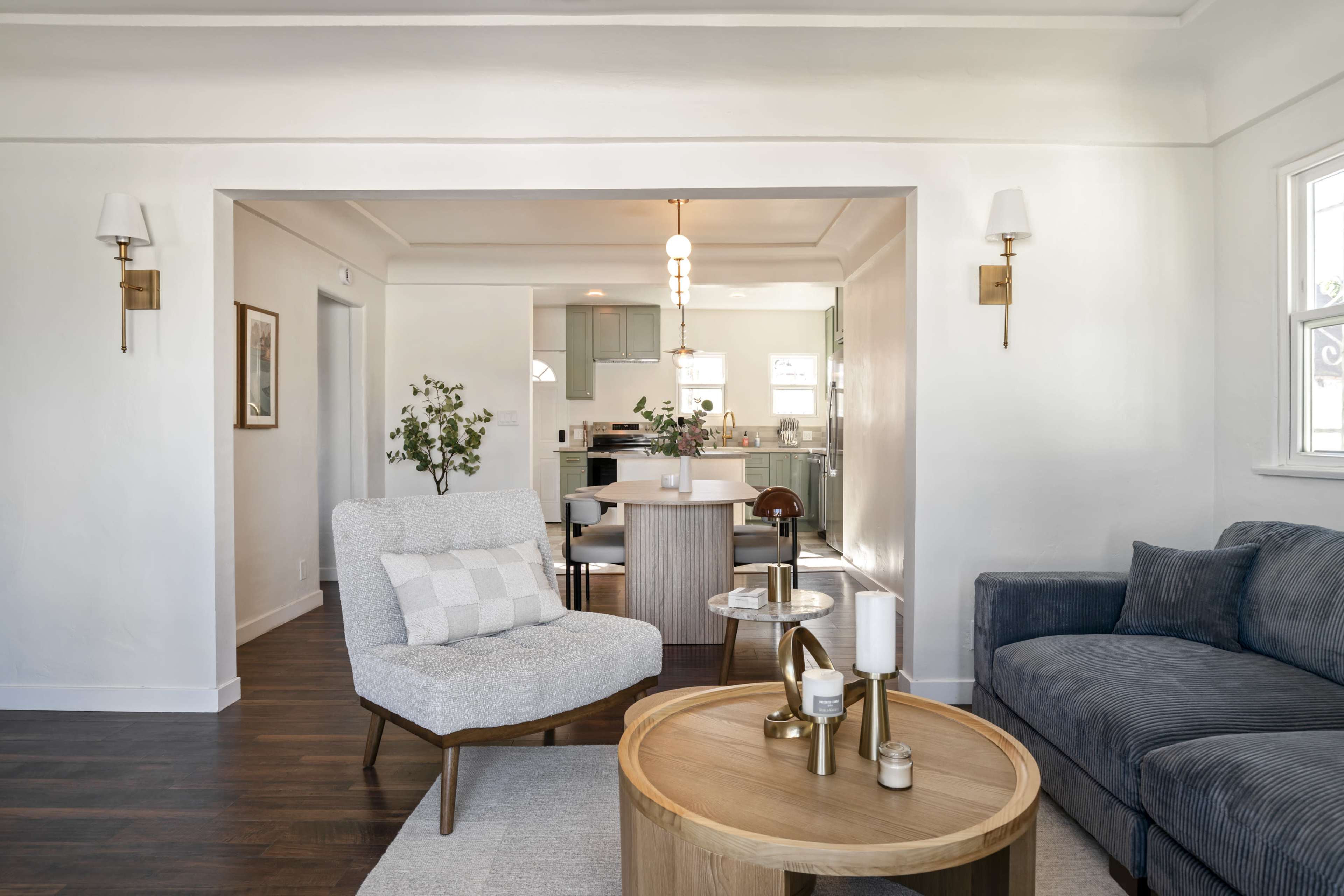 The image shows a modern living room with a comfortable chair, a round coffee table, and a view of a dining area in the background.