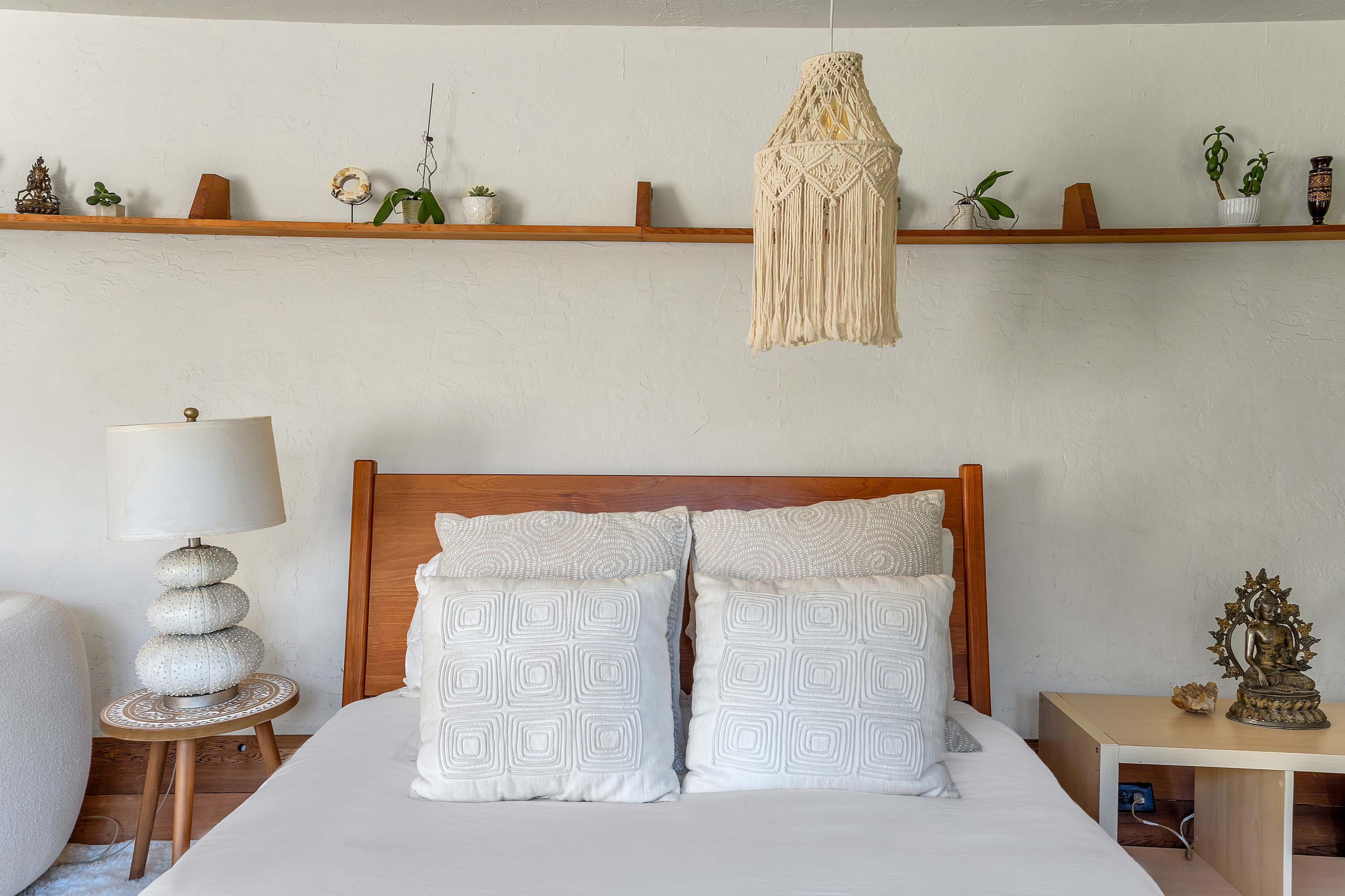 A wooden bed with white bedding and two decorative pillows sits beneath a hanging light fixture, while a shelf above displays small plants and decorative items.