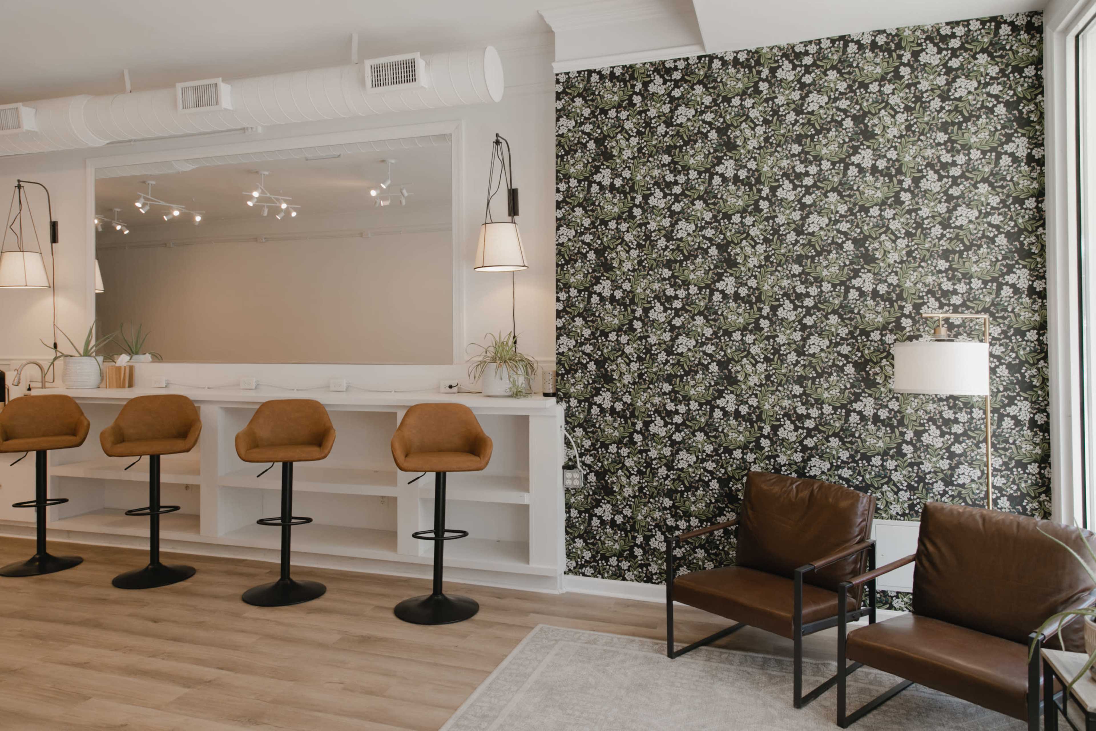A modern interior space with a floral accent wall, a bar area with four tall stools, and two brown leather chairs.