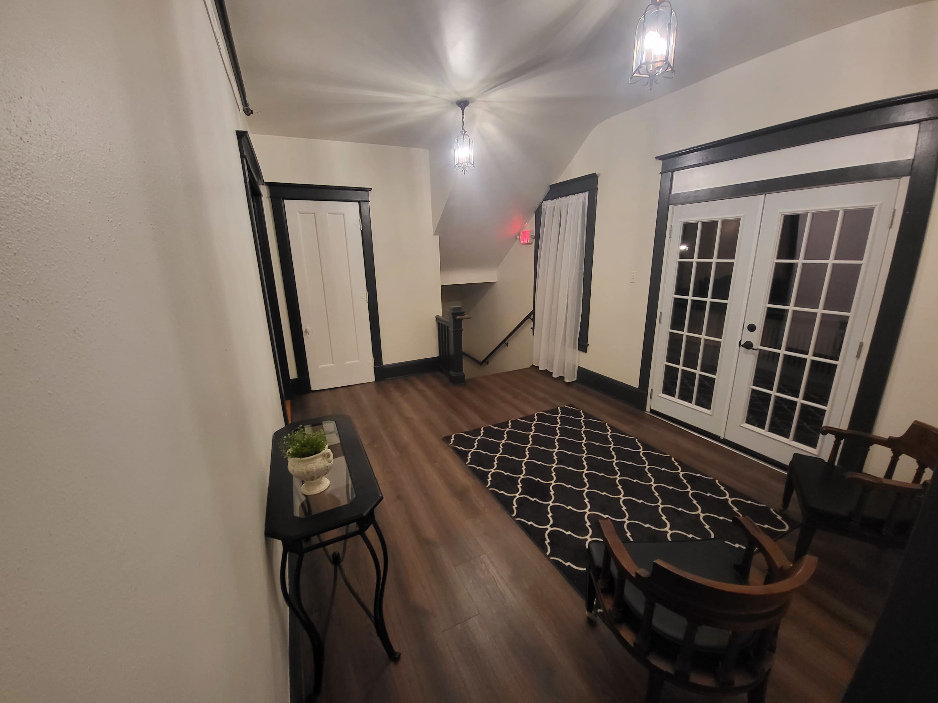 An interior hallway with a black and white patterned rug, two wooden chairs, a small table with a potted plant, and double doors leading to an outdoor area.