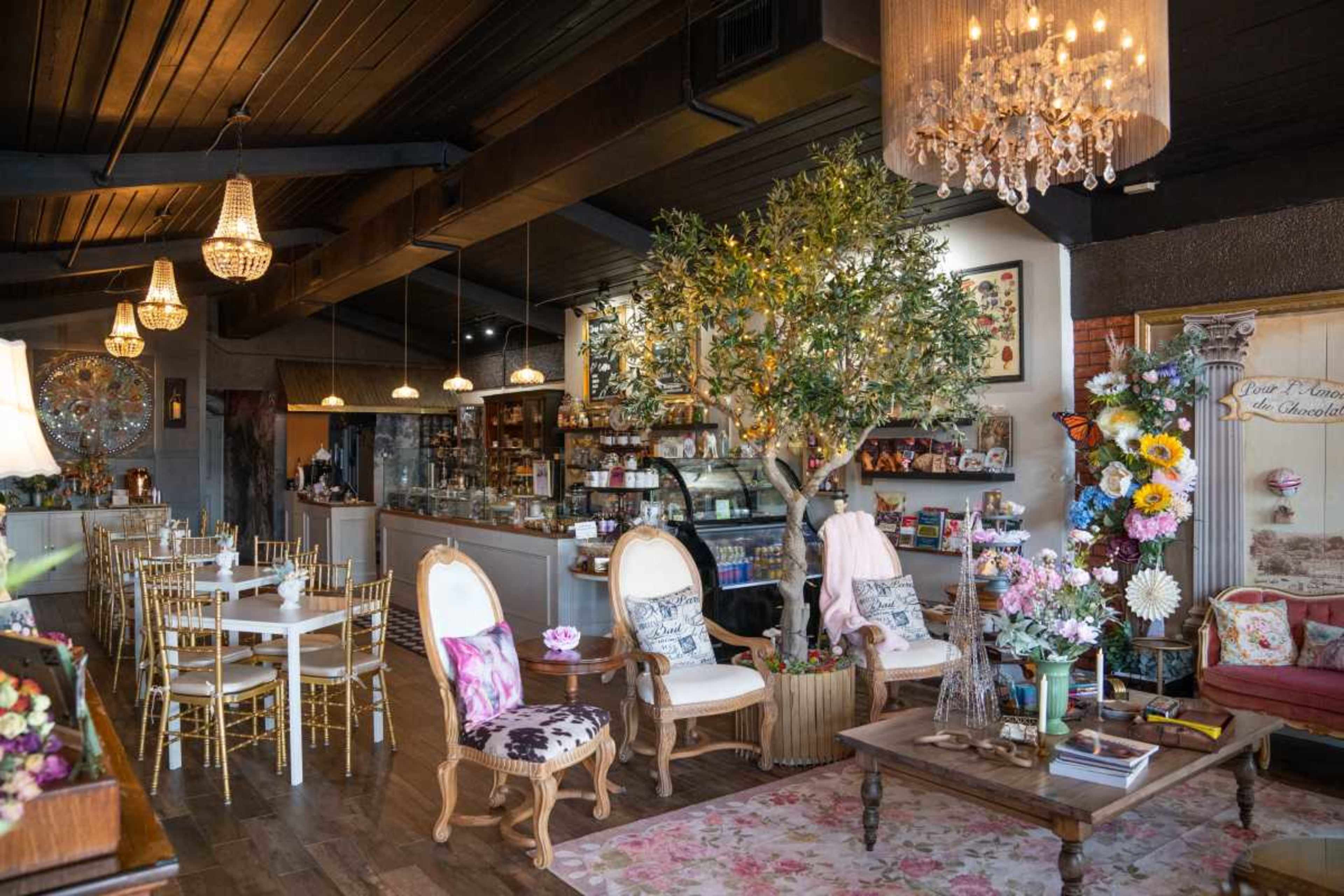 The image shows a cozy café interior with wooden floors, elegant seating, decorative plants, and a display area for food and drinks.