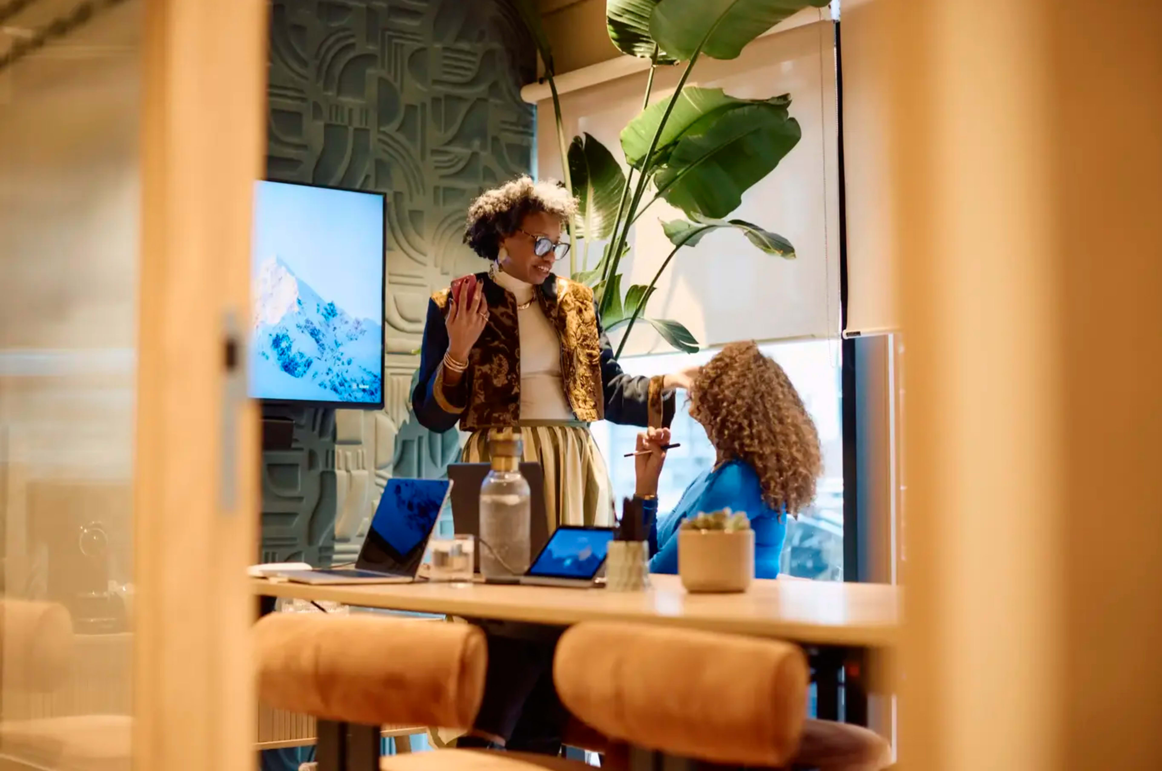 Two women are engaged in conversation in a modern office setting with a large plant and a screen displaying a mountain image in the background.