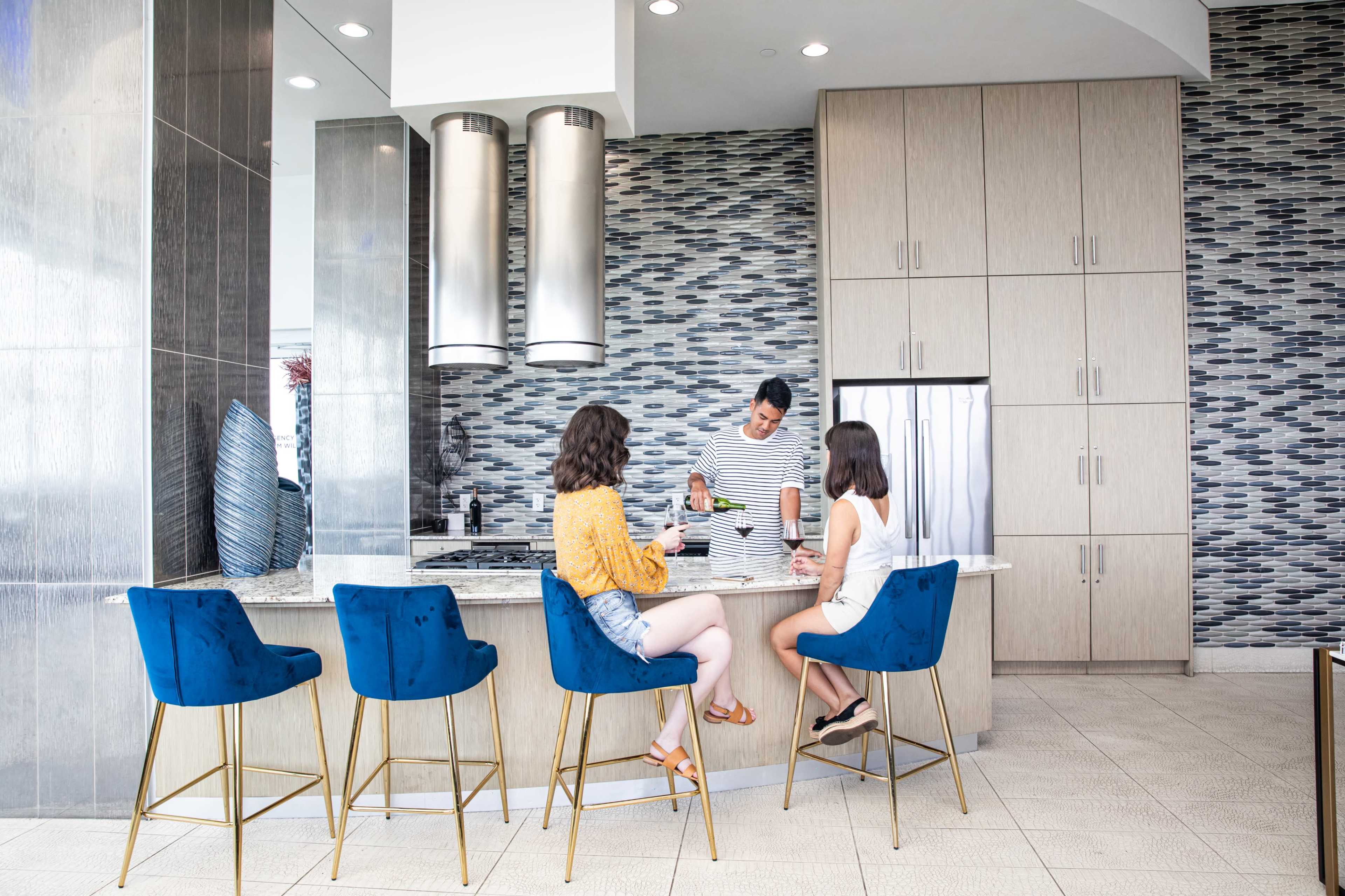 Two women sit at a modern bar counter while a male staff member prepares drinks in a stylish kitchen area.