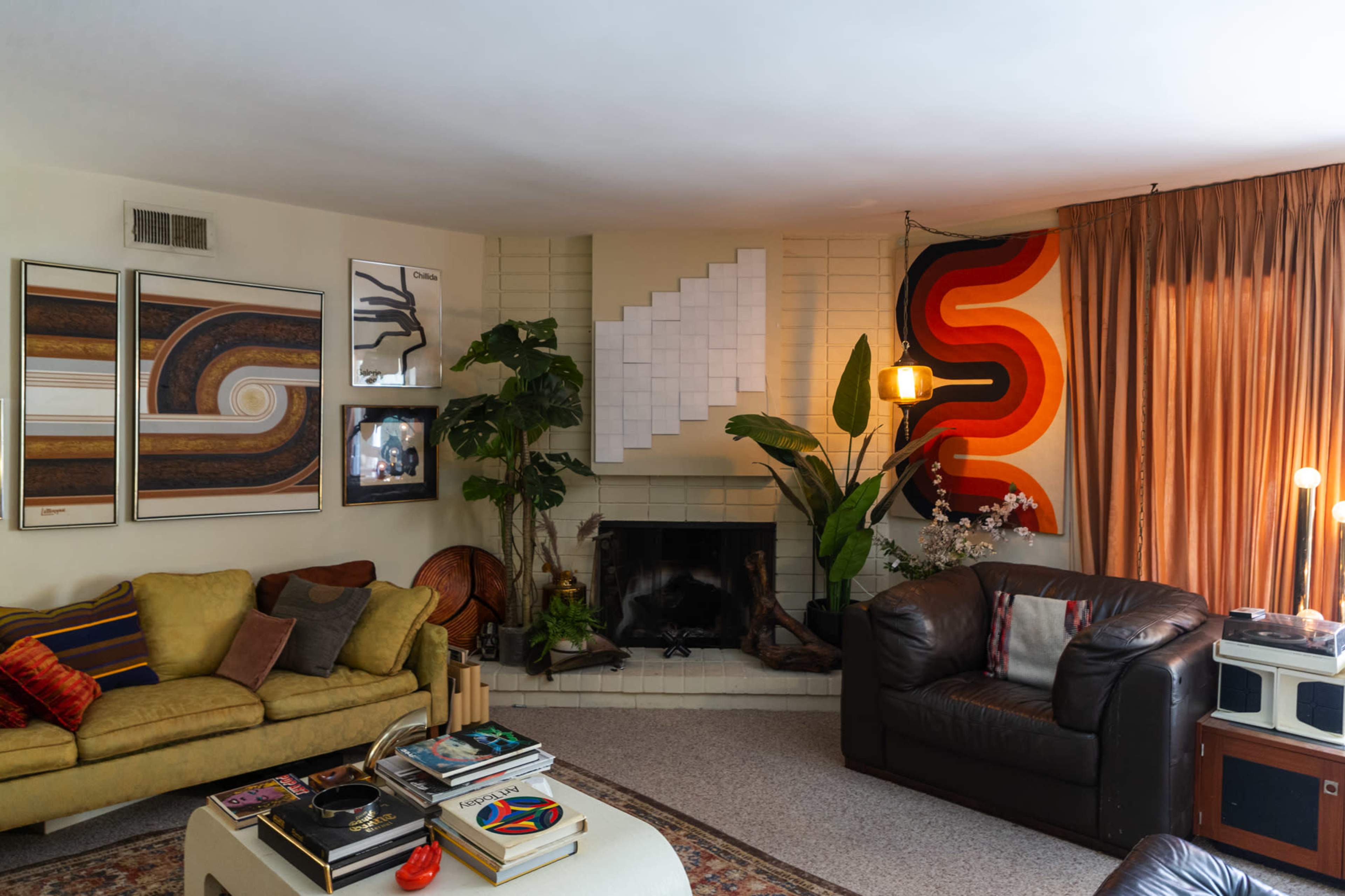 The image shows a cozy living room with a fireplace, decorative plants, and a combination of patterned artwork on the walls.