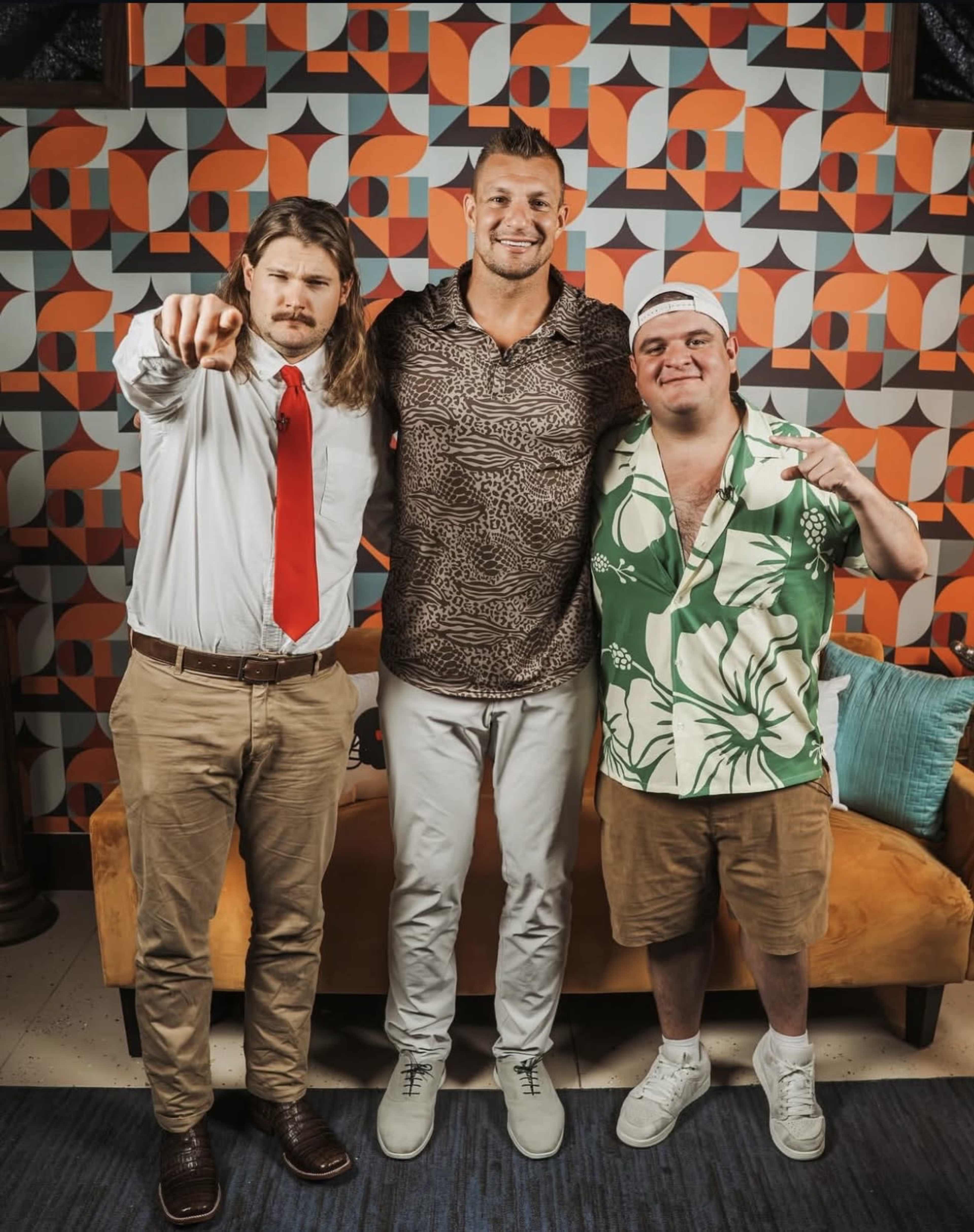 Three men stand together, one wearing a patterned shirt, while the other two are dressed in formal and casual attire, against a backdrop of geometric patterned wallpaper.