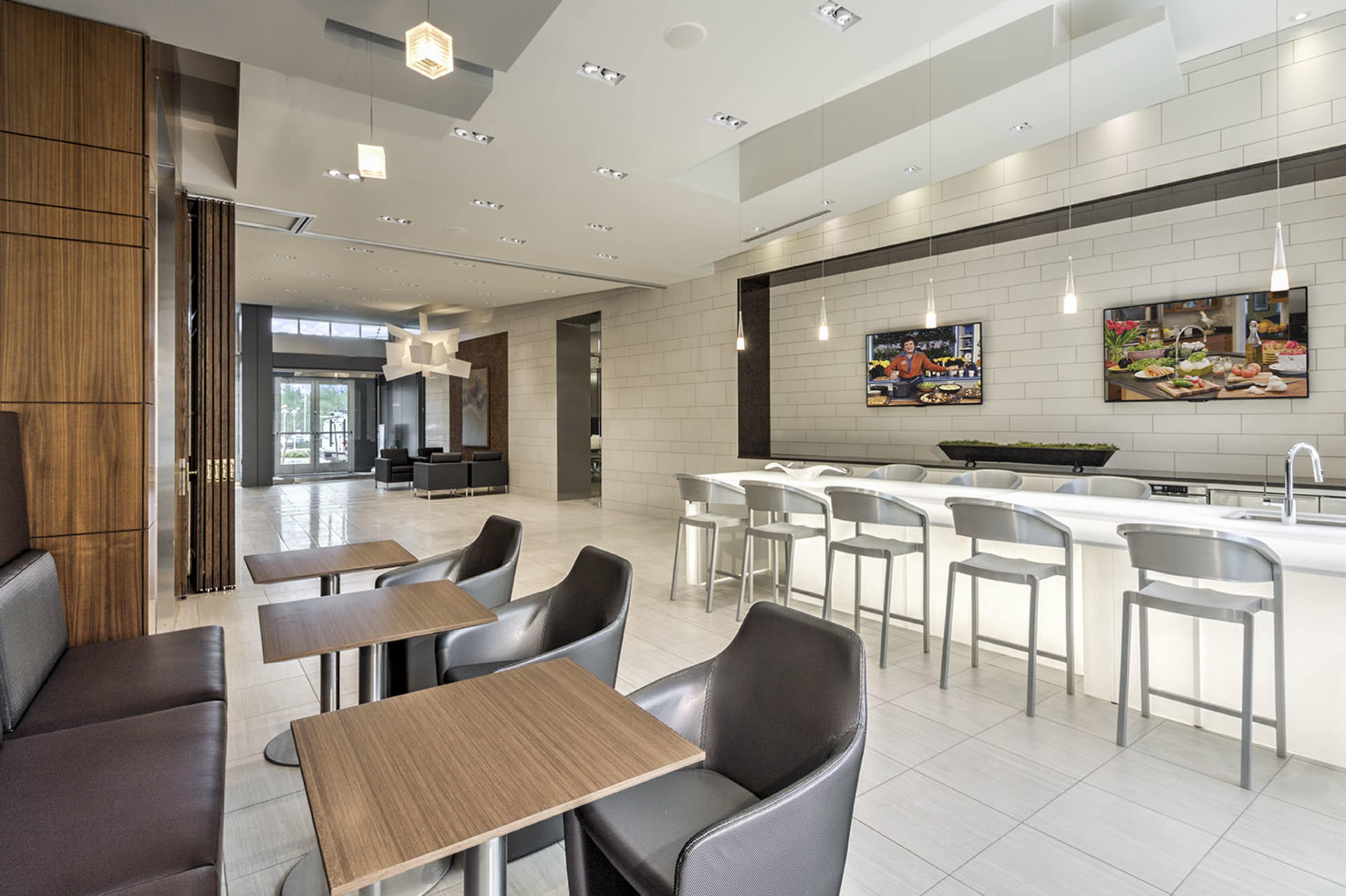A modern lobby area with a seating arrangement featuring small wooden tables and sleek chairs, alongside a well-lit bar counter displaying two screens.