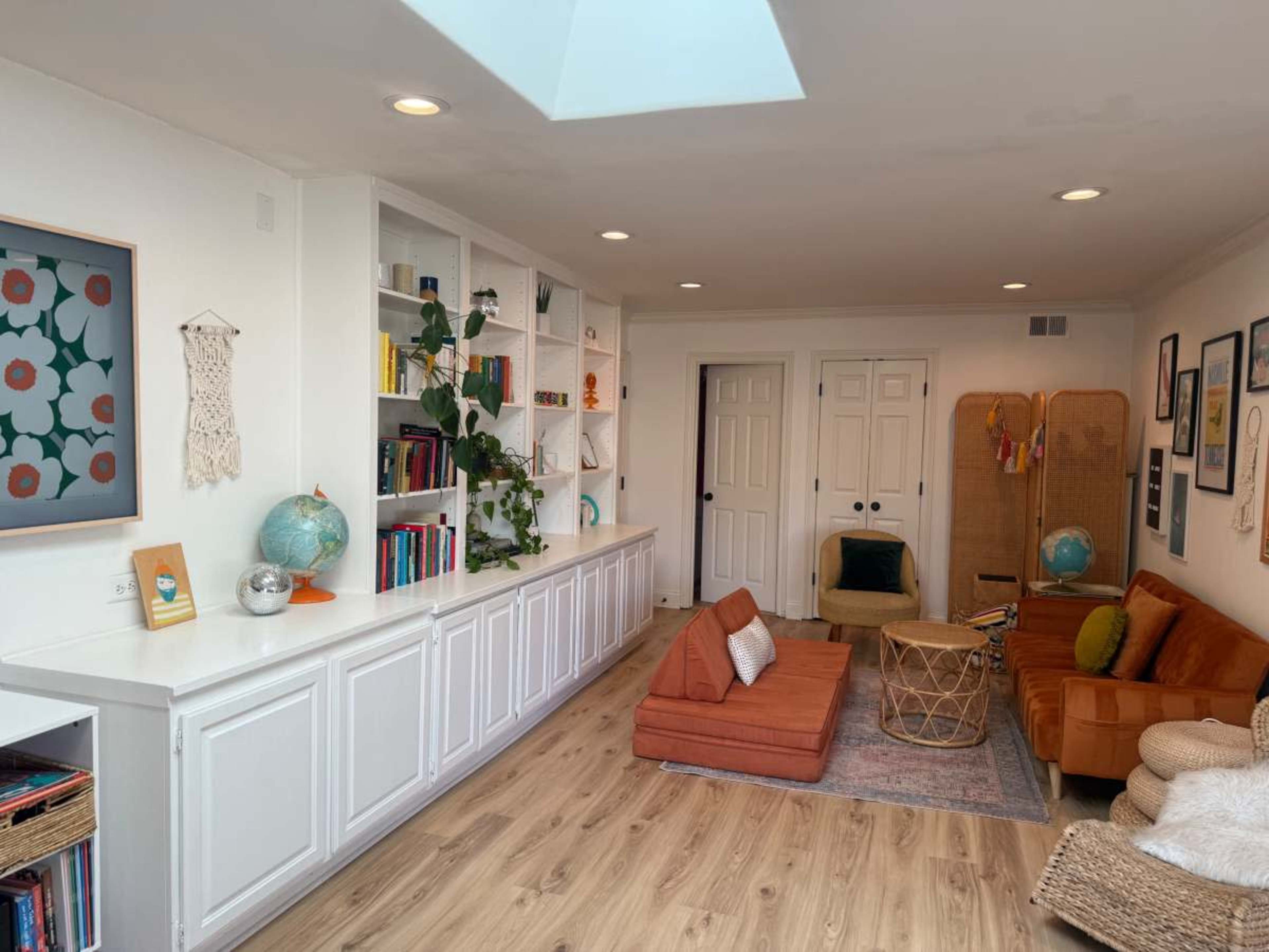 The image shows a cozy living room with orange couches, a large bookshelf filled with books, and a skylight illuminating the space.