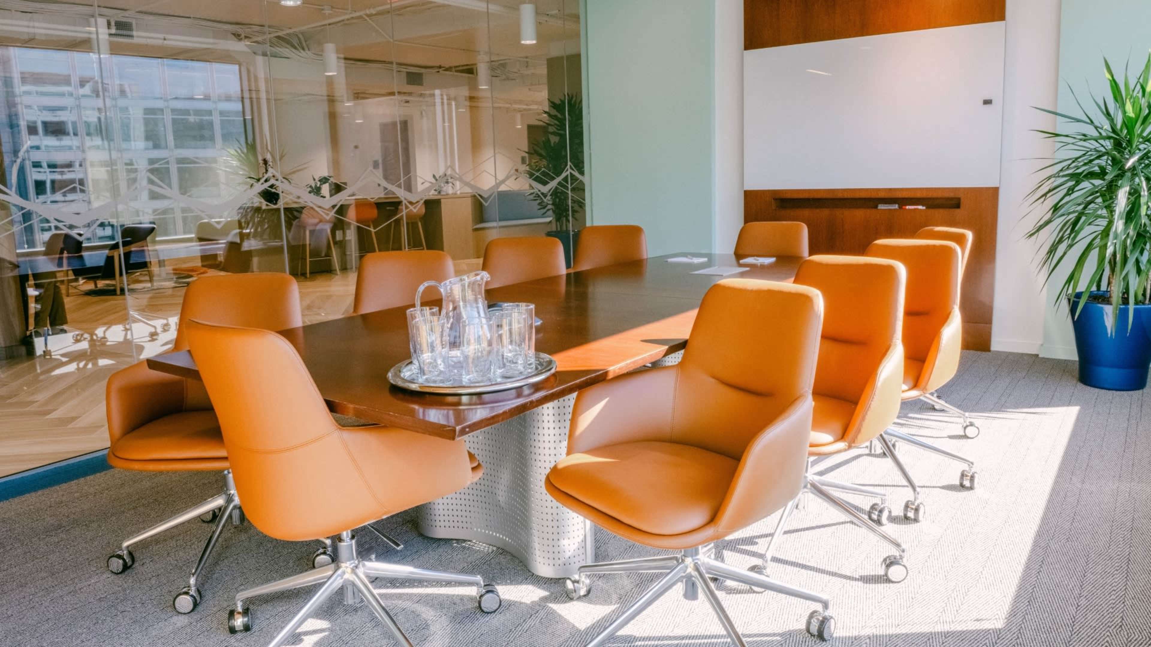 A modern conference room features a rectangular wooden table surrounded by eight orange leather chairs and glass water pitchers on a tray.