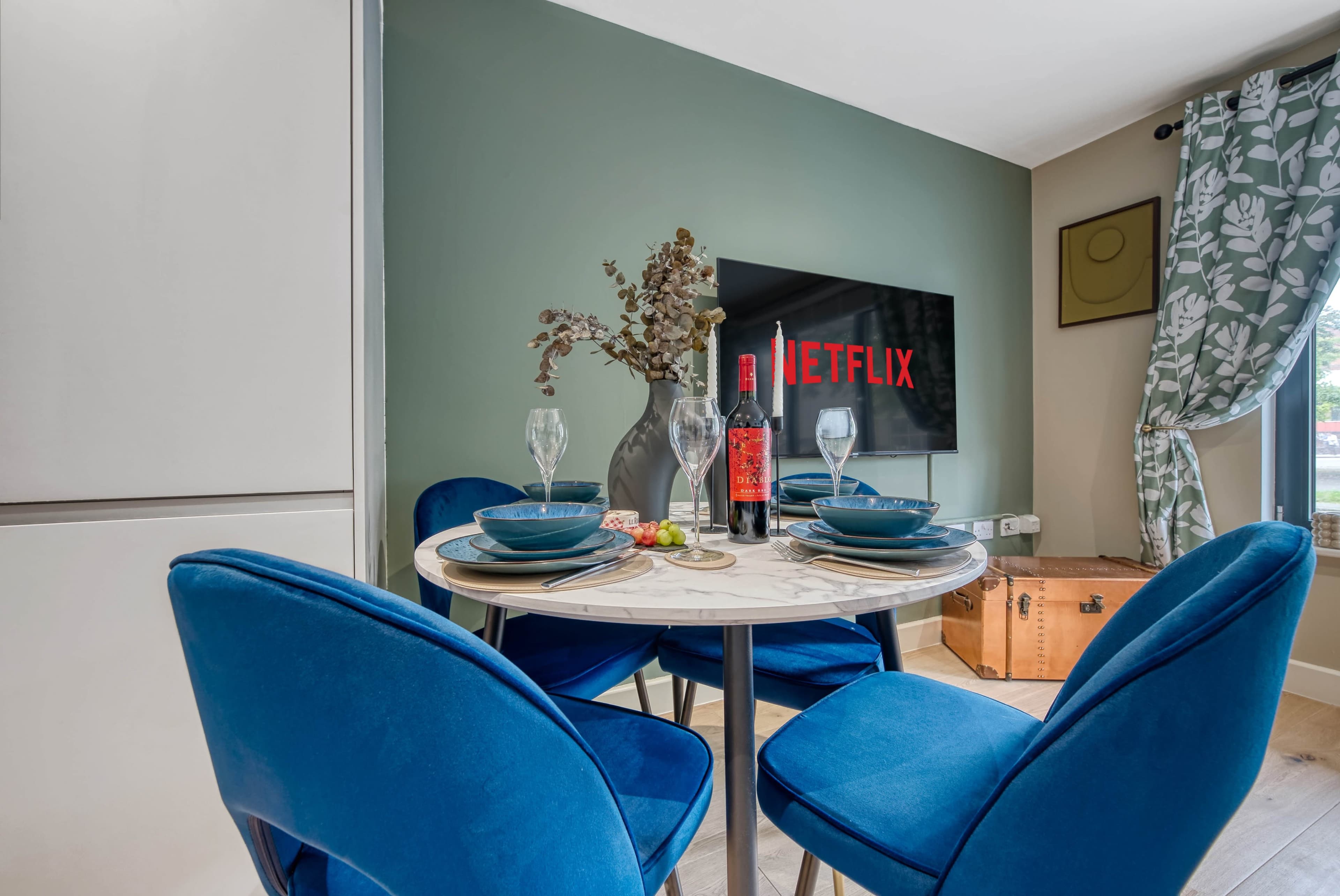 A small dining area features a round table set with blue chairs, a decorative bottle, and a television displaying the Netflix logo.