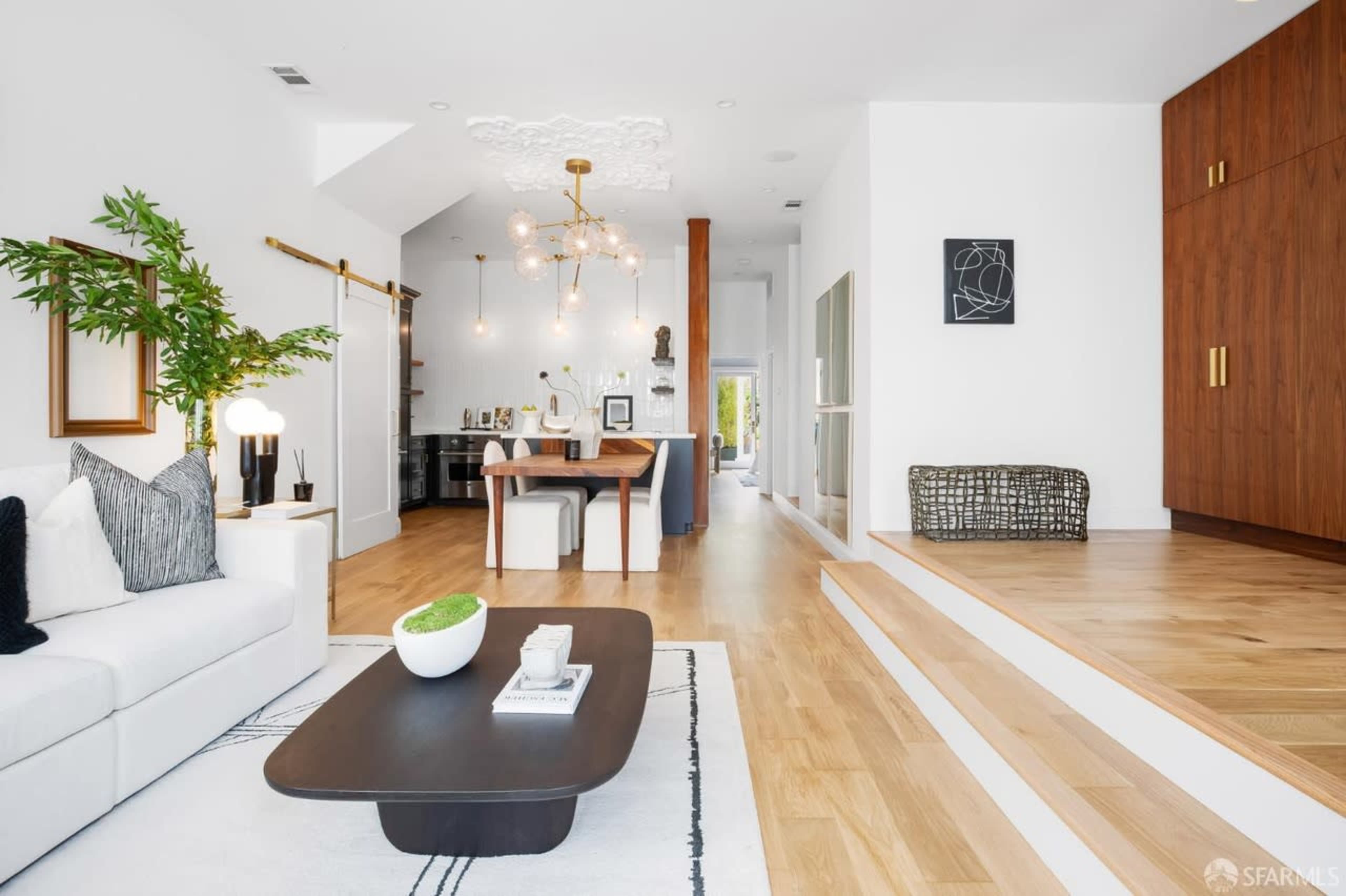 The image shows a modern living space with a white sofa, a dark coffee table, a dining area with a wooden table, and steps leading to a different area of the room.