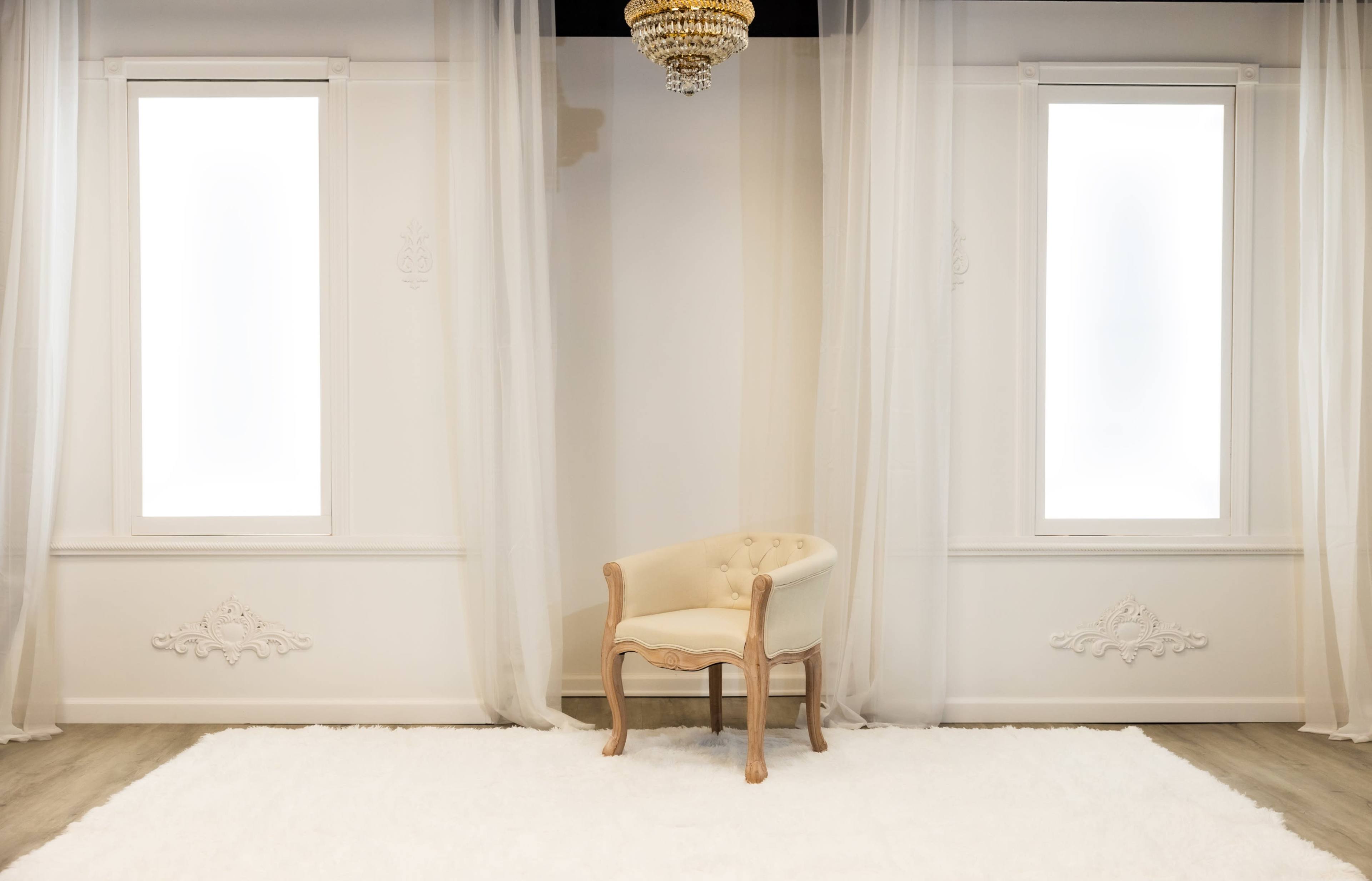 A single upholstered chair is positioned in the center of a brightly lit room with large windows and sheer curtains.